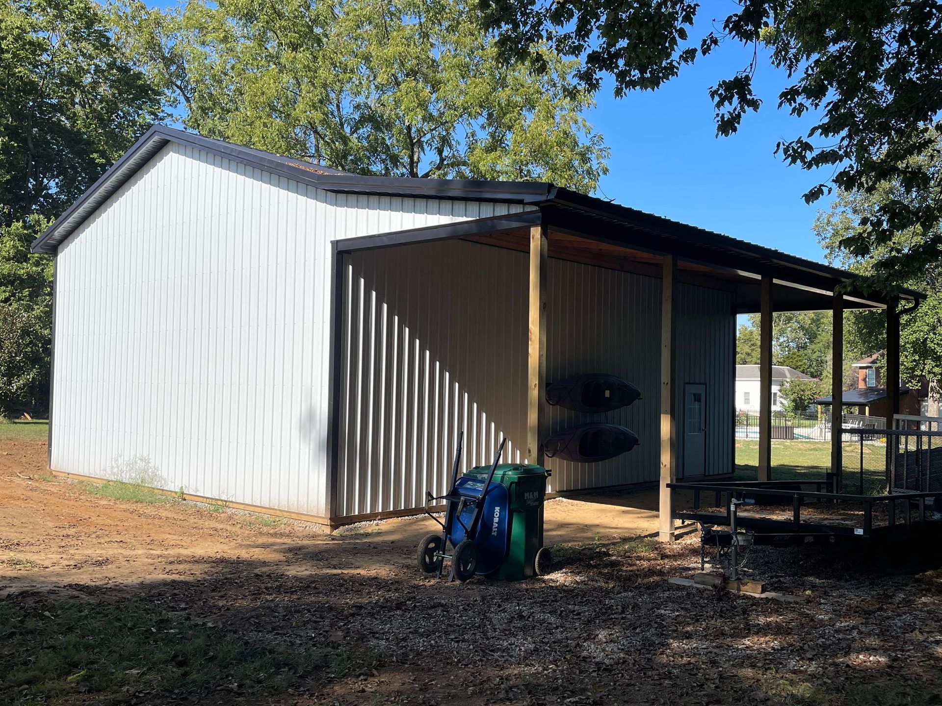 A white metal shed with a black roof and a covered area, kayaks inside, and a trailer nearby.