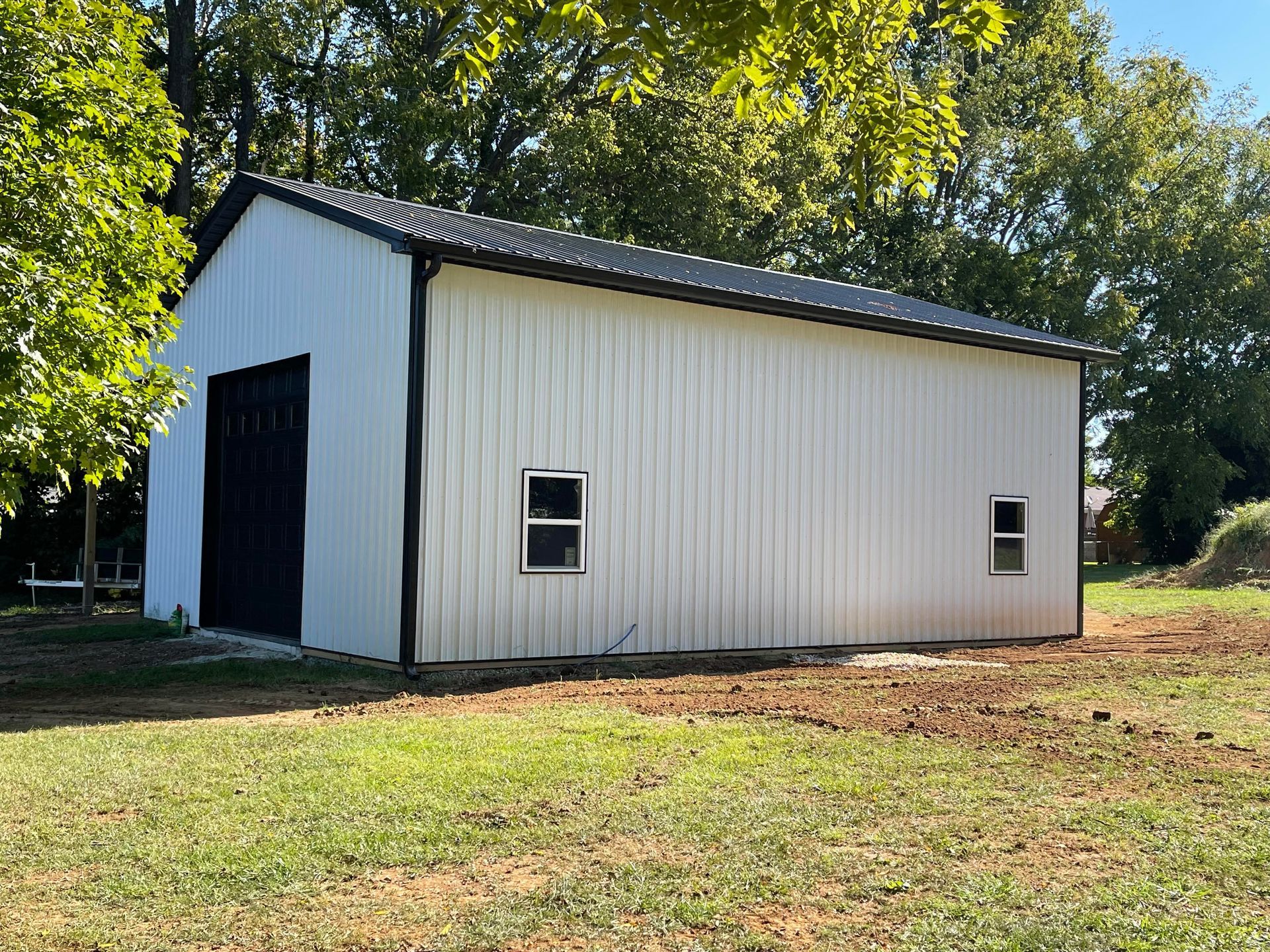 White metal barn with a black roof and large door, two small windows. Set in a grassy yard with trees in the background.