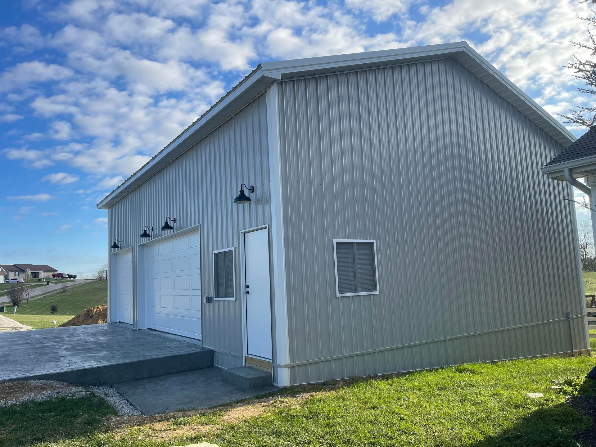 A silver metal garage with a white door and garage doors, set on a concrete slab against a blue sky and grassy yard.