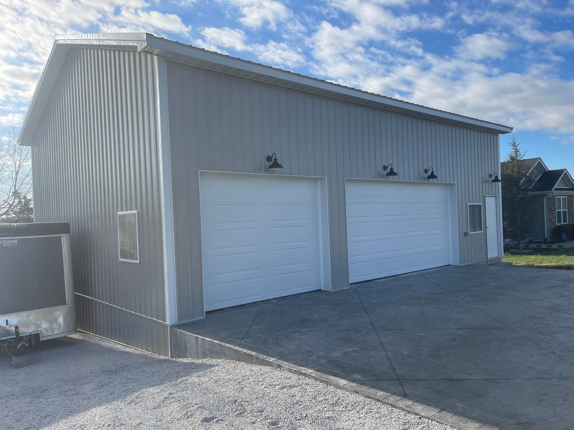 Gray metal garage with two white garage doors and side entry door under a blue sky. Gravel in the foreground.