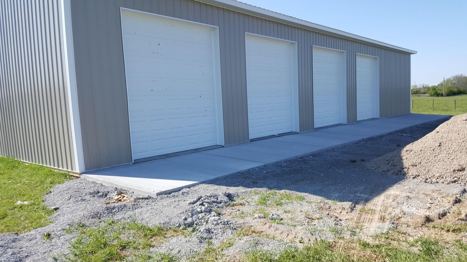 A grey metal building with three white garage doors, a concrete walkway, and a gravel bed in a grassy outdoor setting.