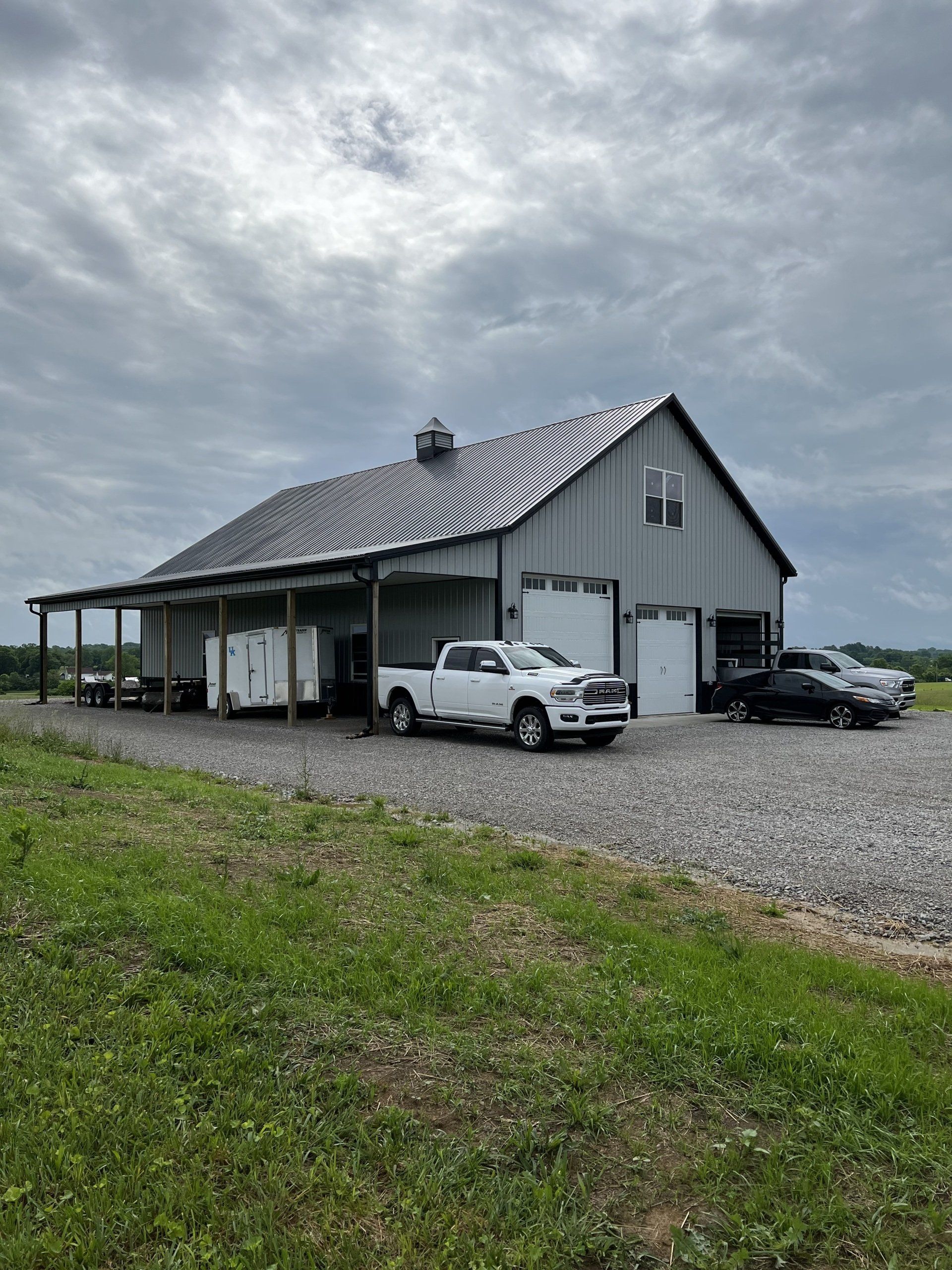 A white barn with a covered porch, gravel drive, and gray metal roof. A white truck, a trailer, and two cars are parked outside.