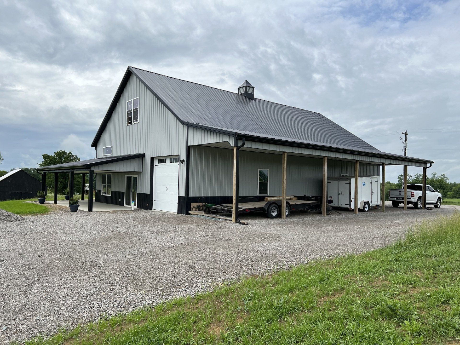 Gray barn with a black roof and covered porches, parked vehicles on a gravel drive, cloudy sky.