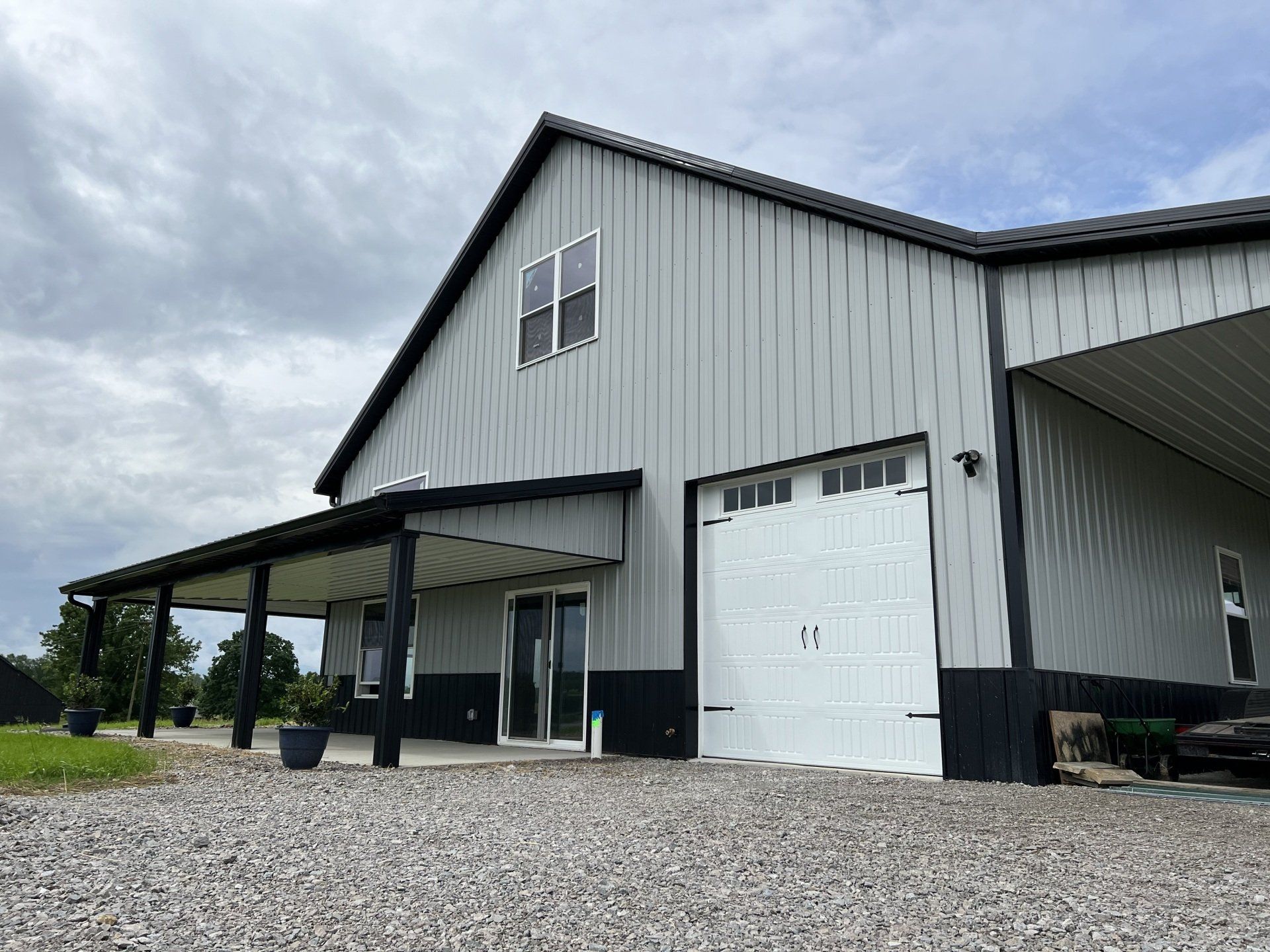 A gray metal building with a white garage door and a covered porch. The trim and the lower portion of the exterior are painted black.
