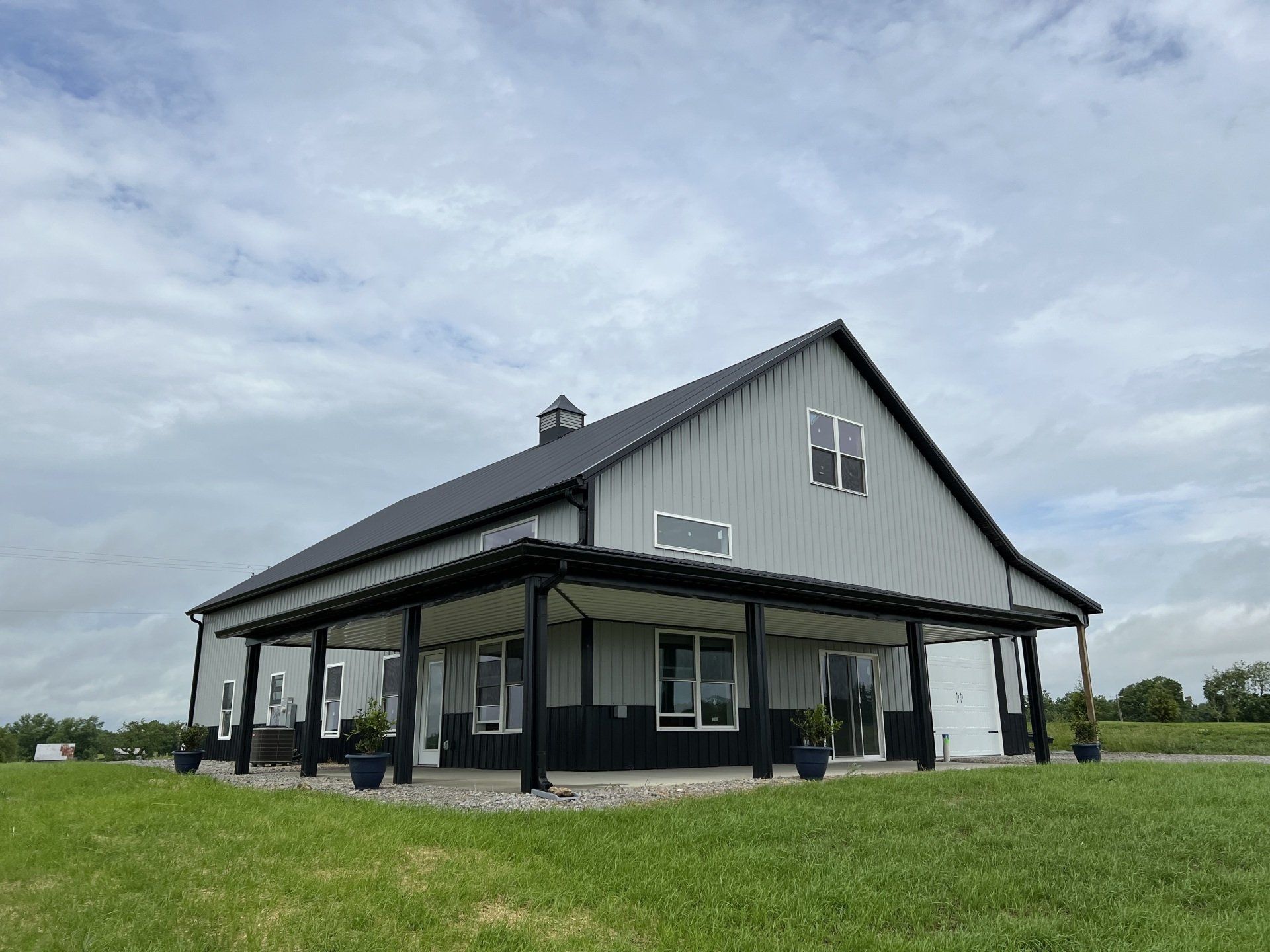 Gray barn-style building with a black roof and wraparound porch, standing in a green field under a cloudy sky.