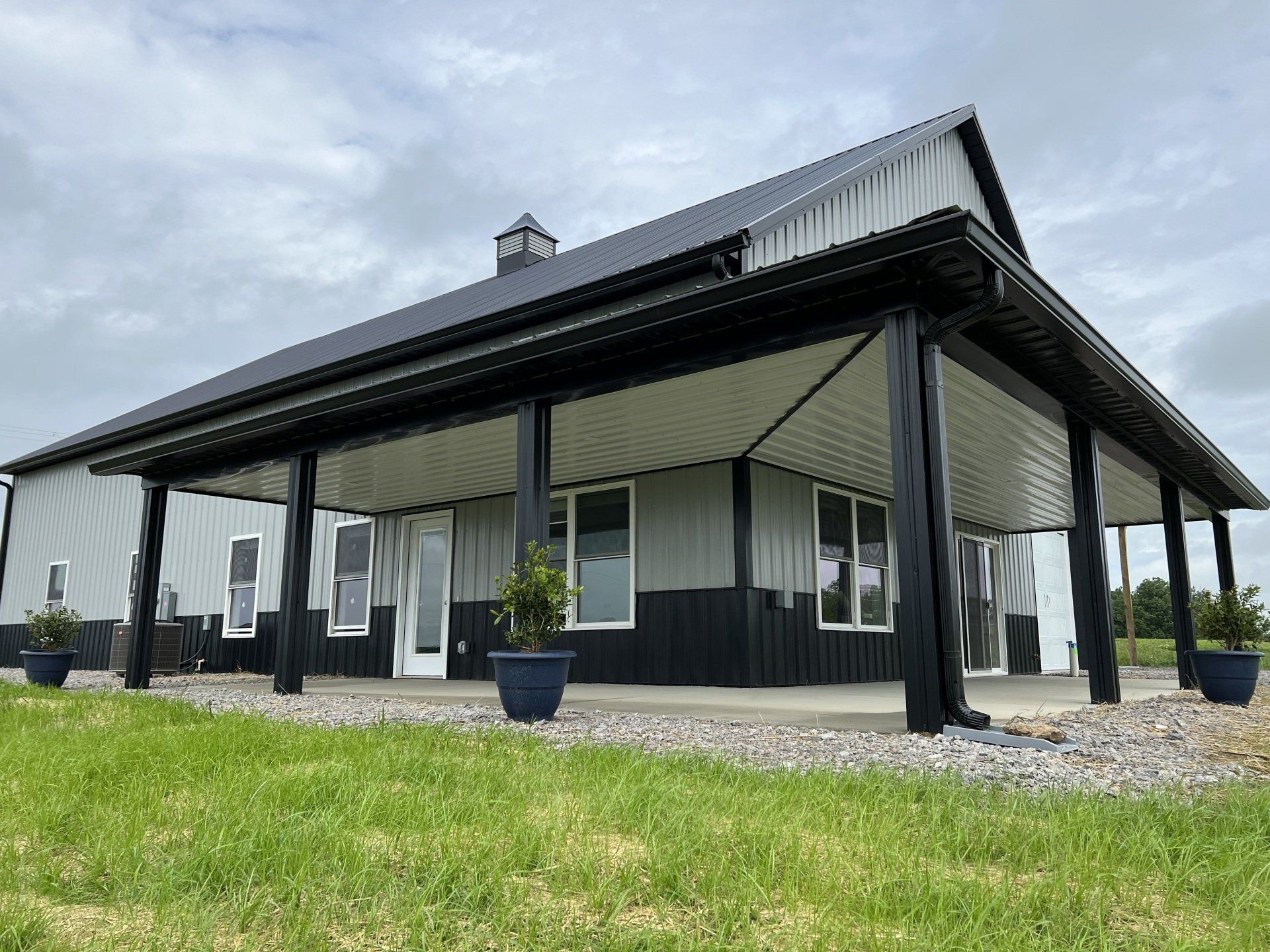 A modern gray and black barn with a large covered porch, overlooking a grassy field under a cloudy sky.