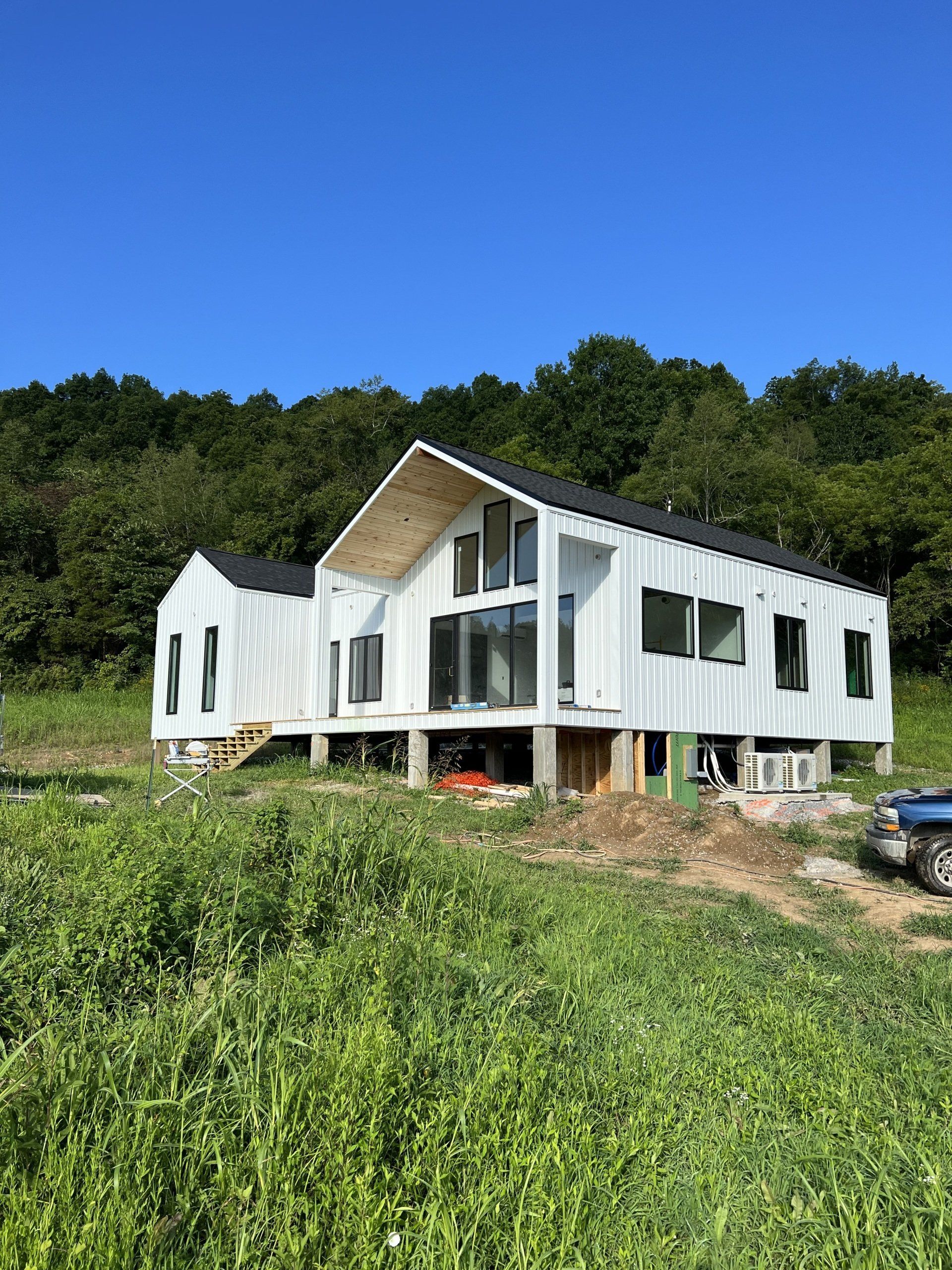 White modern house under construction with black roof and windows, set against a hillside of trees and blue sky.