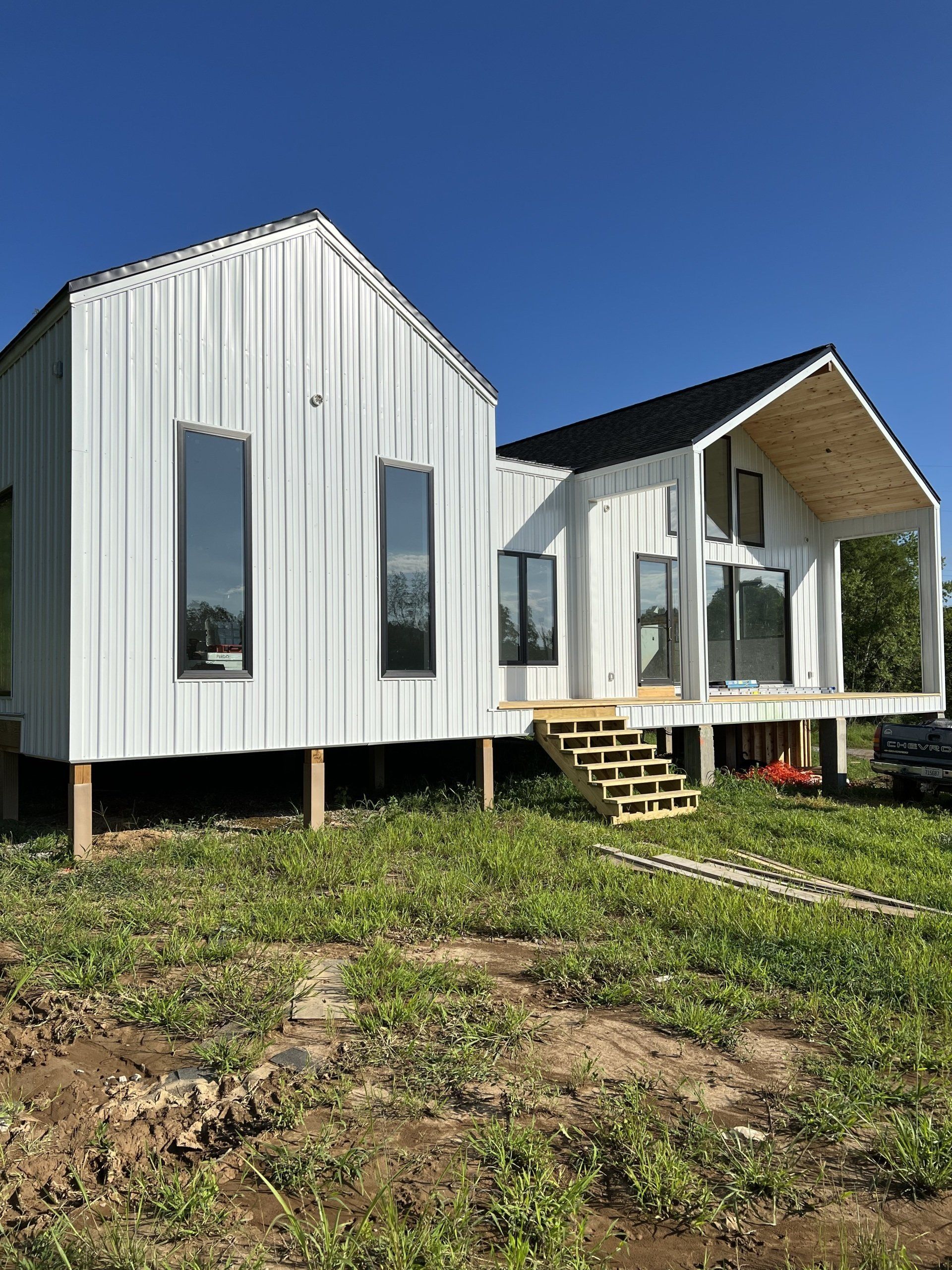 White modern house with angled roof, black windows, and wooden porch, set on wooden stilts in a field with green grass under a blue sky.