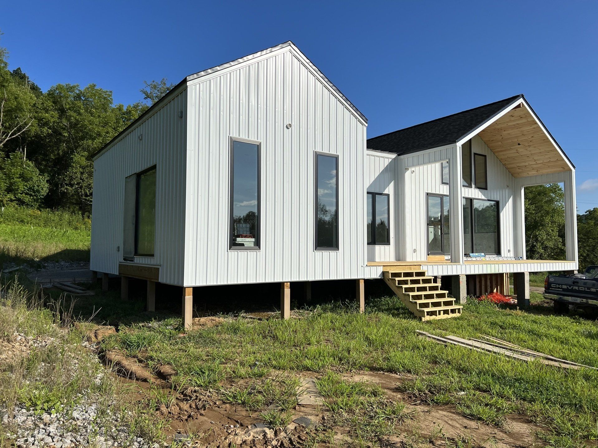 Modern white house with two connected gabled sections, elevated on wooden supports.  Tall, narrow windows, black roof, and a porch are visible.