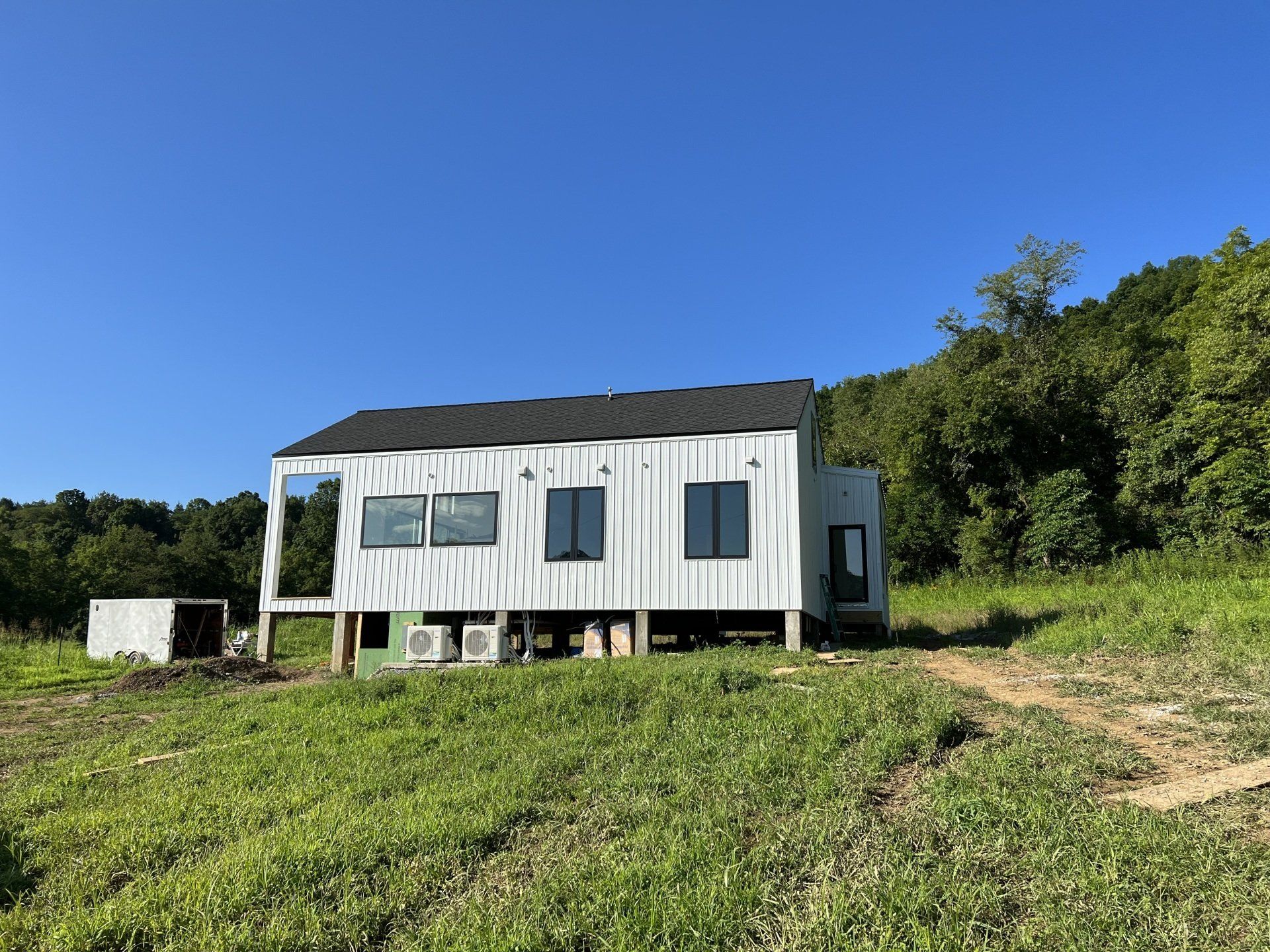 White cabin on stilts in a grassy field, with a dark roof and blue sky. Trees in the background.