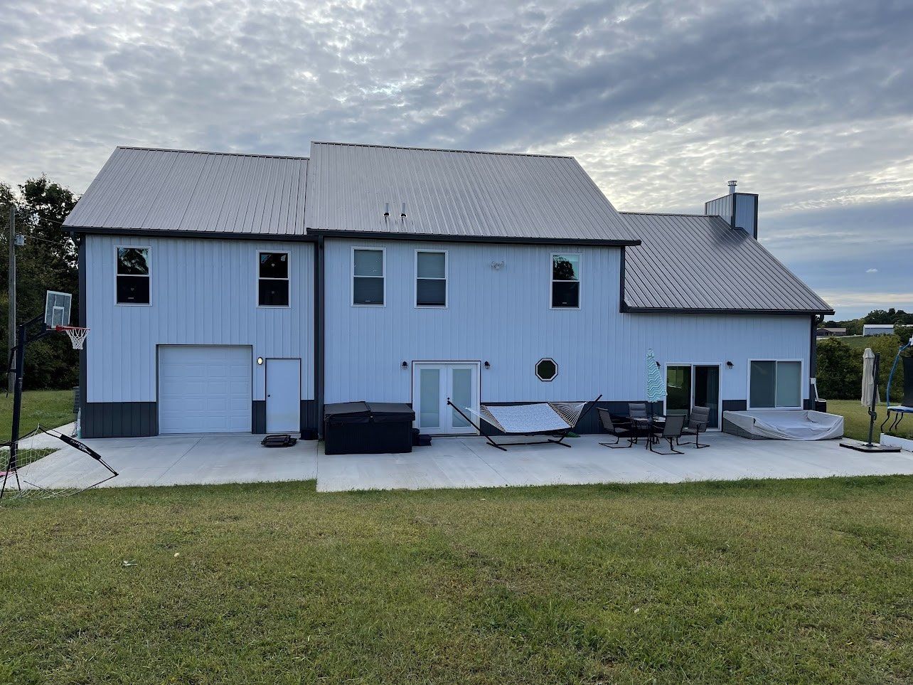 Back of a light blue house with a silver metal roof, a concrete patio, and a grassy lawn under a cloudy sky.