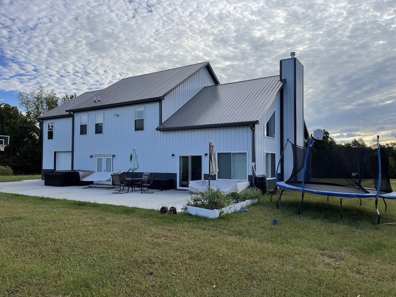 A two-story white house with a black metal roof and chimney. A trampoline sits in the backyard.