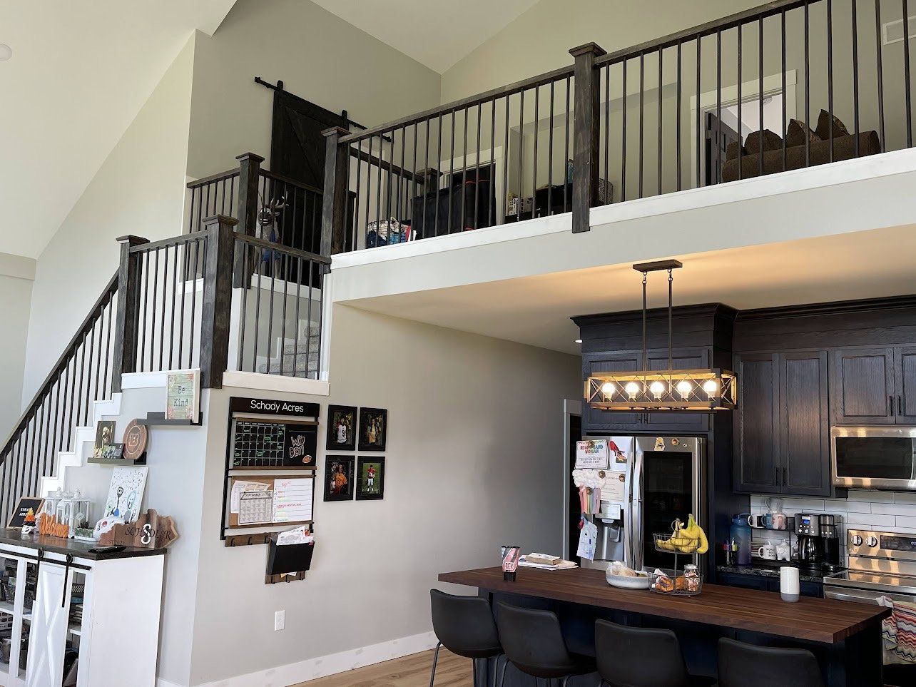 A modern kitchen with a loft area and staircase. Dark cabinets, a wooden island with stools, and a stainless steel refrigerator are visible.