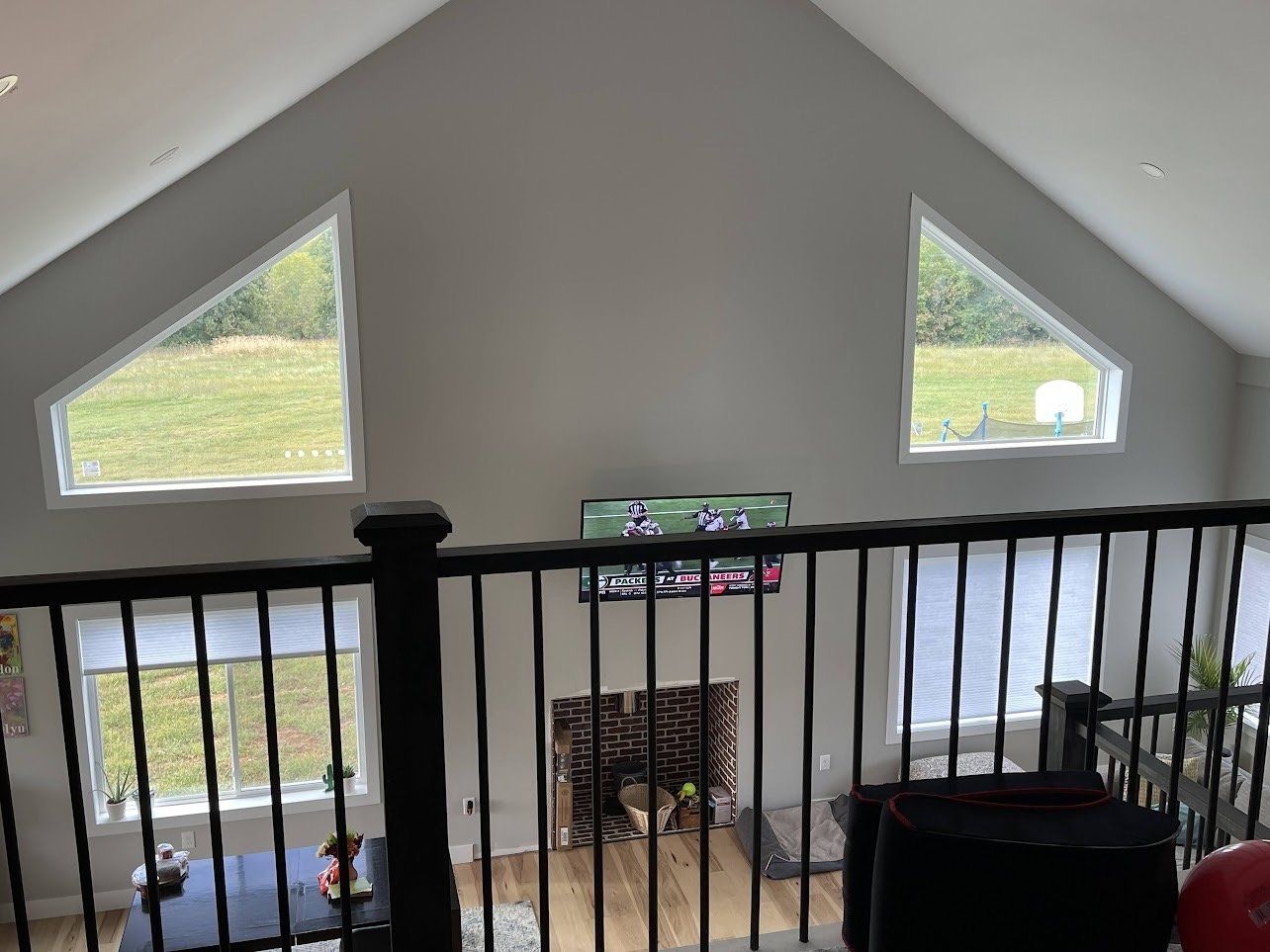 View from a balcony overlooking a living room with a fireplace, TV, and triangular windows in a high ceiling. Black railing.