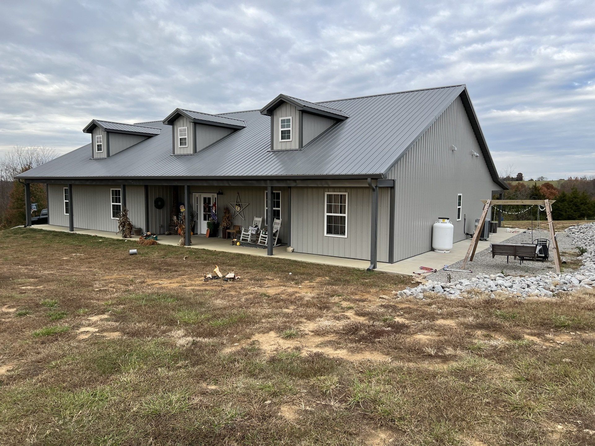 Gray metal-sided house with a porch, three dormers, and a metal roof, set on a grassy lot with a cloudy sky.