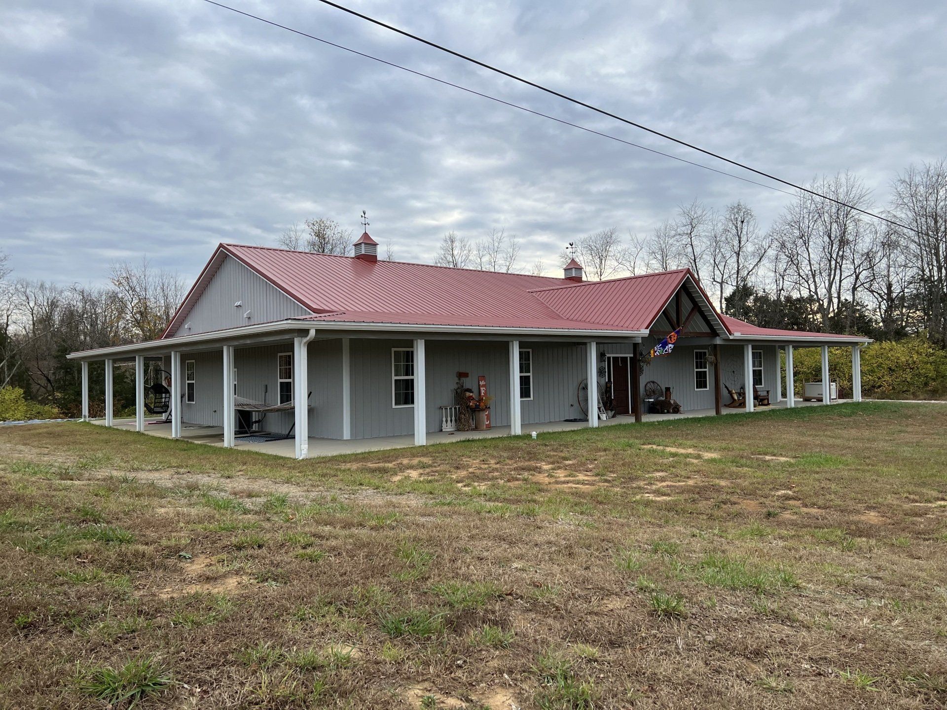 A gray house with a red roof, porch, and white columns, set on a grassy lot under an overcast sky.