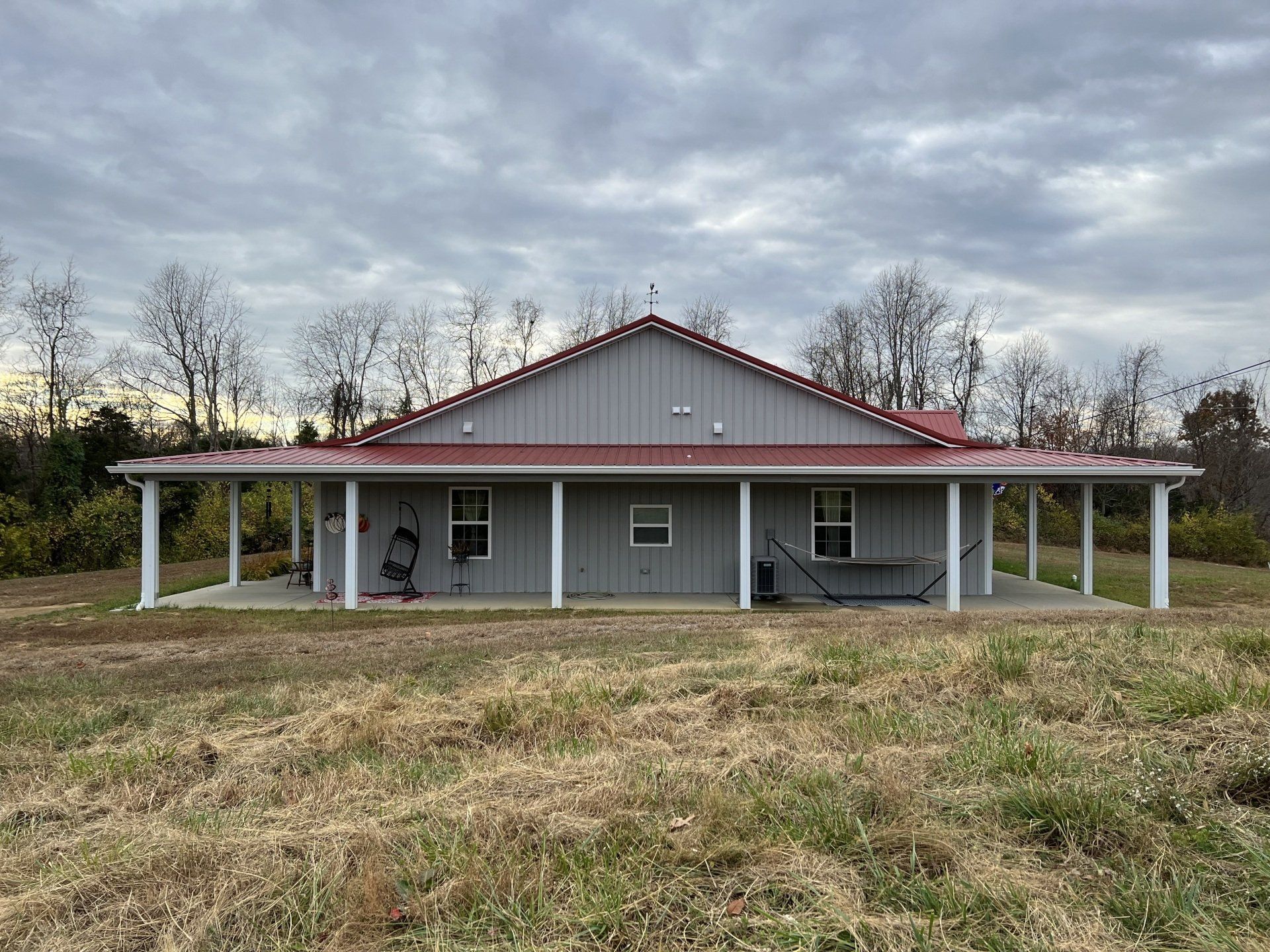 A gray house with a red roof and covered porch stands in a field under a cloudy sky.