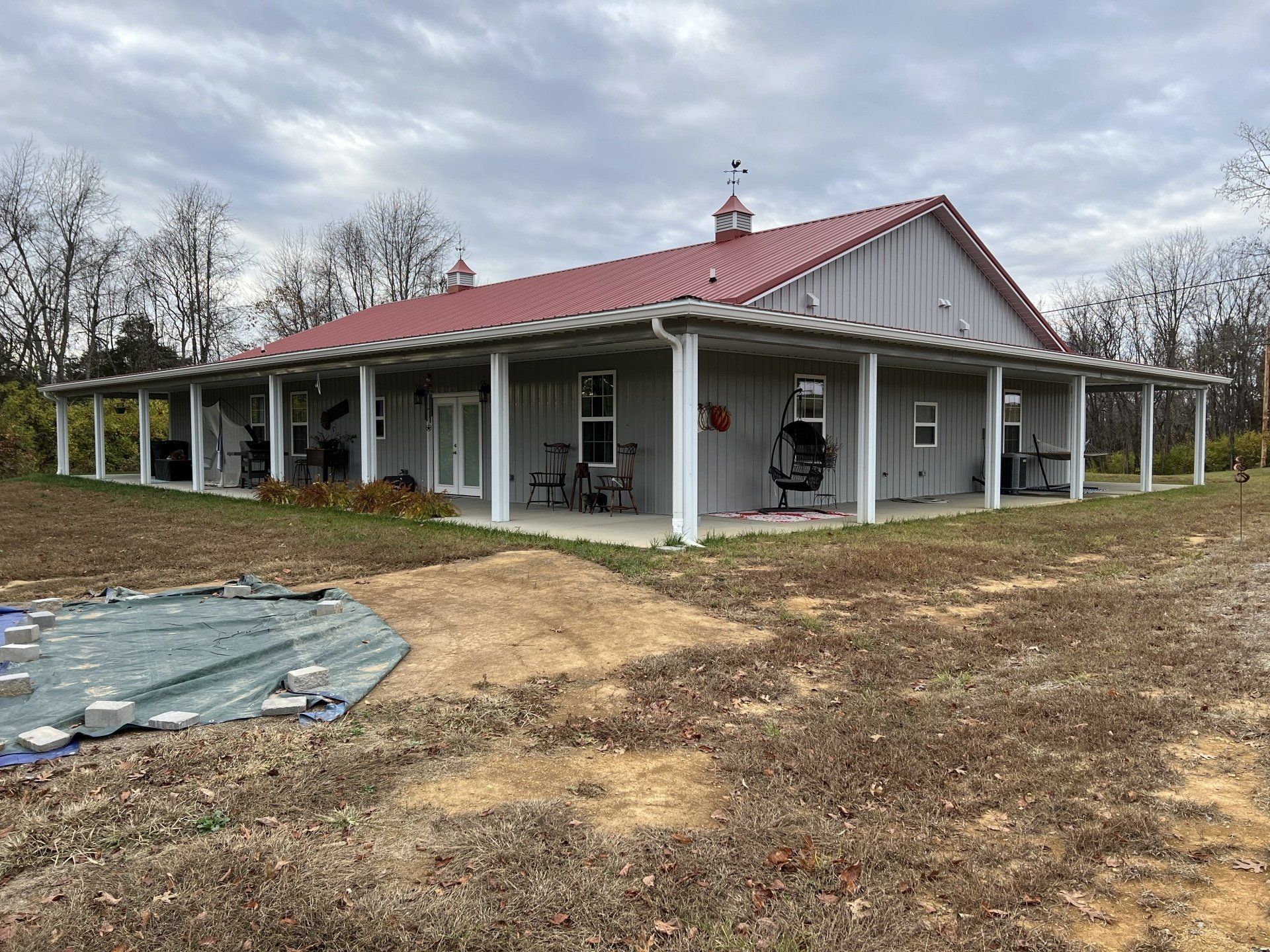 A gray barn-style house with a red roof and a wrap-around porch. The house is set in a grassy yard under a cloudy sky.