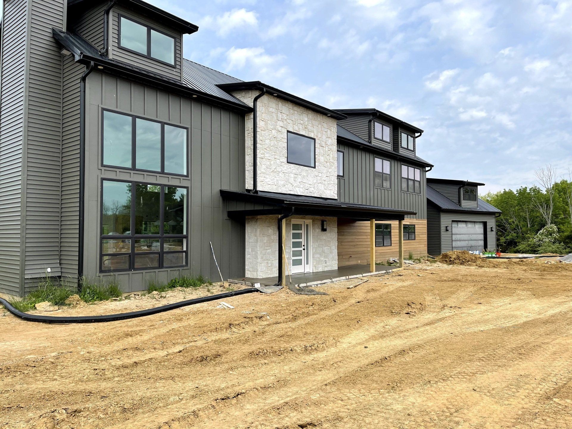 Modern two-story house under construction with dark gray siding and a light brick accent wall. Sandy ground in front.