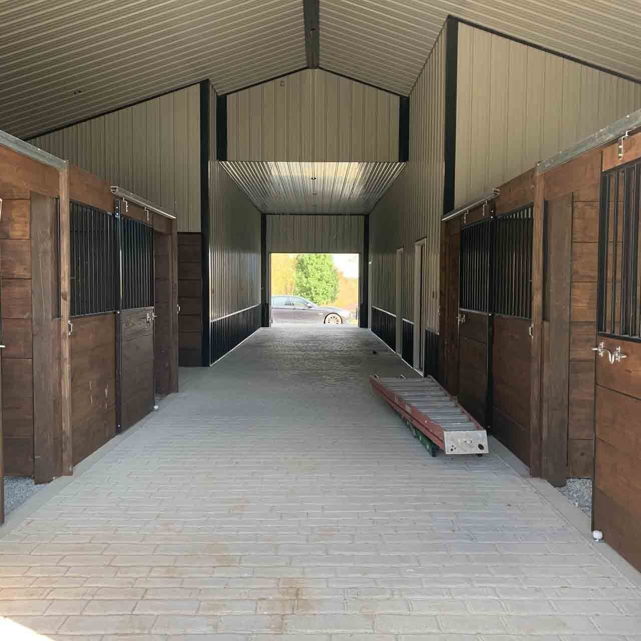 Interior view of a horse stable with wooden stalls lining the sides of a long corridor. Sunlight streams from the open end.