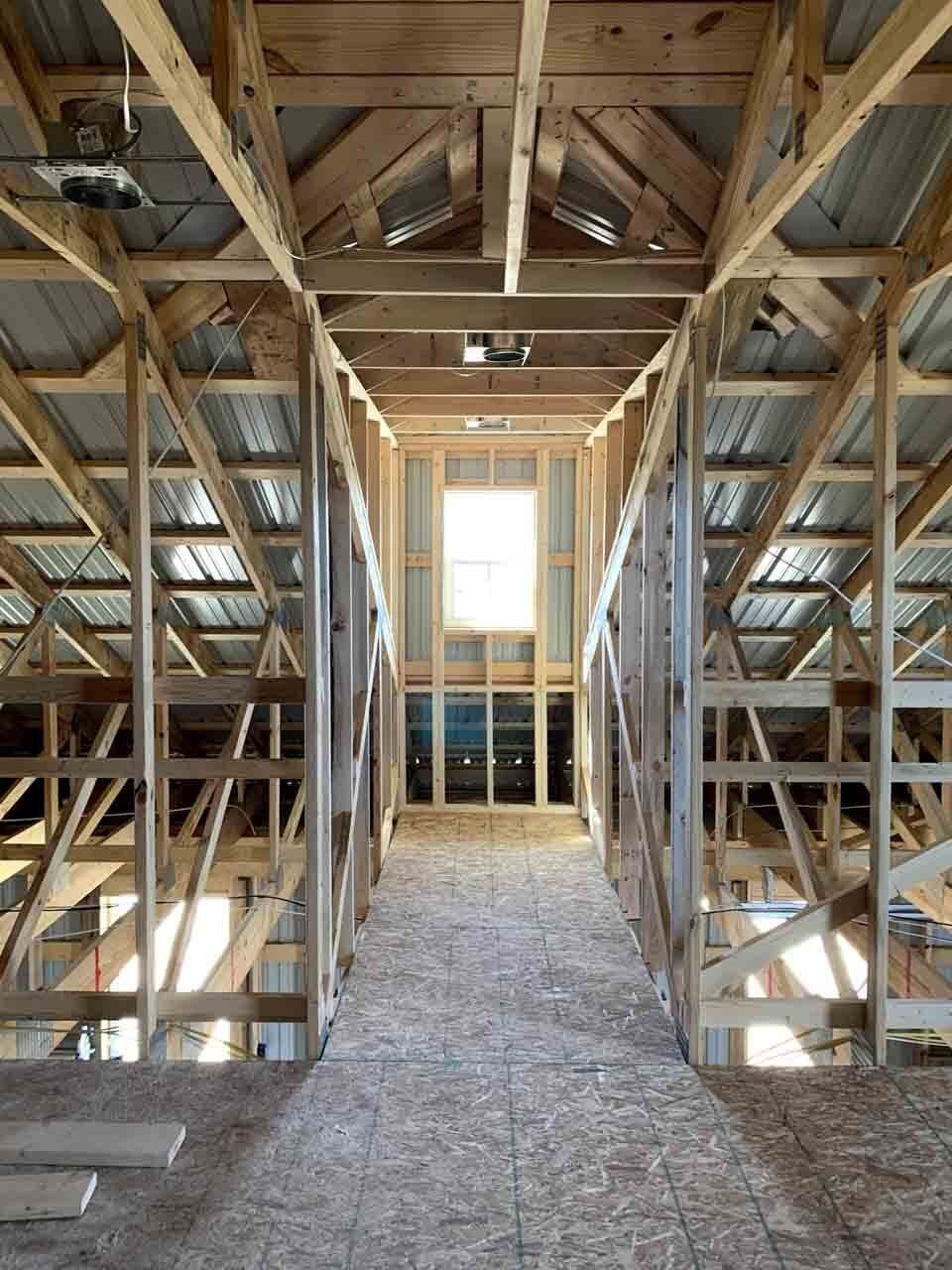 Interior view of a building under construction, showcasing wooden framing and a central hallway leading to a window. Light streams in, illuminating the unfinished structure.
