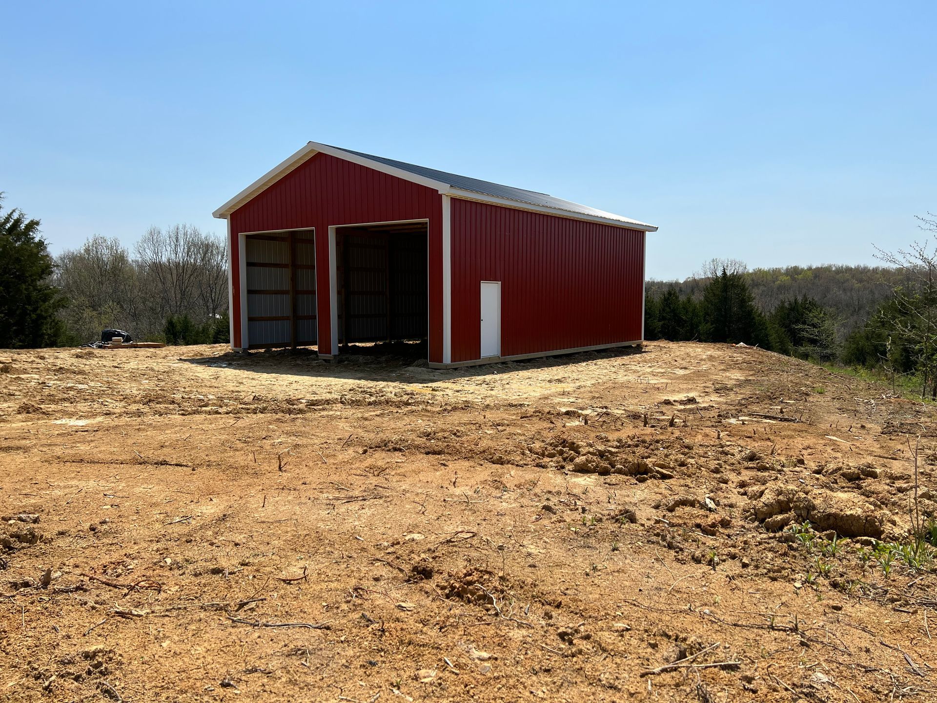 Red metal shed with open garage doors and a white door sits on a dirt lot under a blue sky.