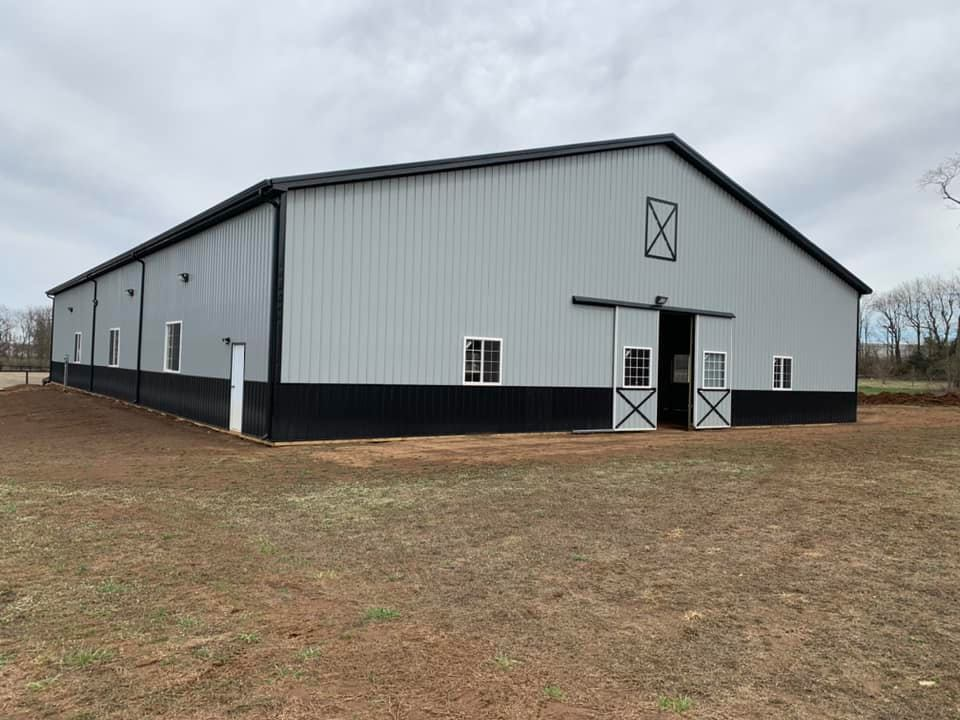 A large, gray and black metal barn with white window and doors, set in a grassy field under a cloudy sky.