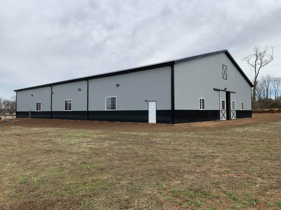 Large gray and black barn with a black roof, white doors and windows, in a grassy field under a cloudy sky.