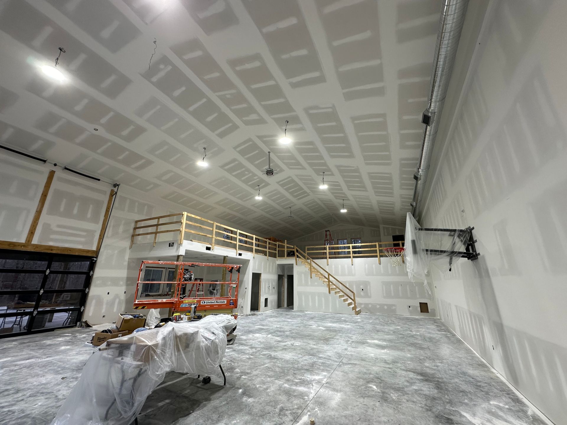Construction interior with drywall-covered walls and ceiling, a second-floor mezzanine, and a lift; the space is empty.