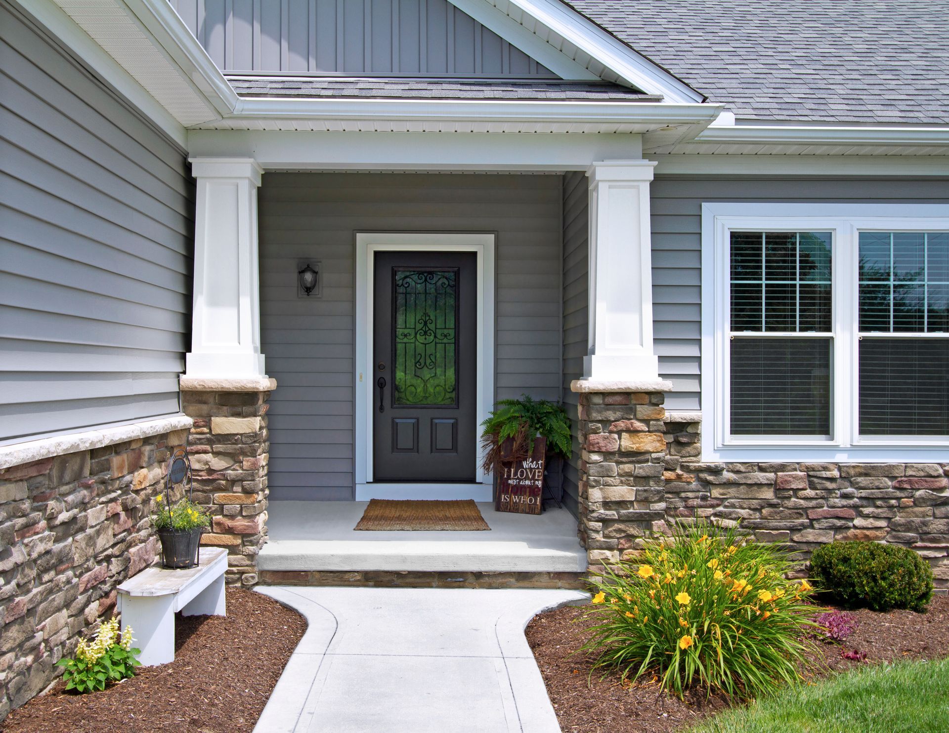 A gray house entrance with a black door, white columns, and a stone facade. A concrete path leads to the front door.