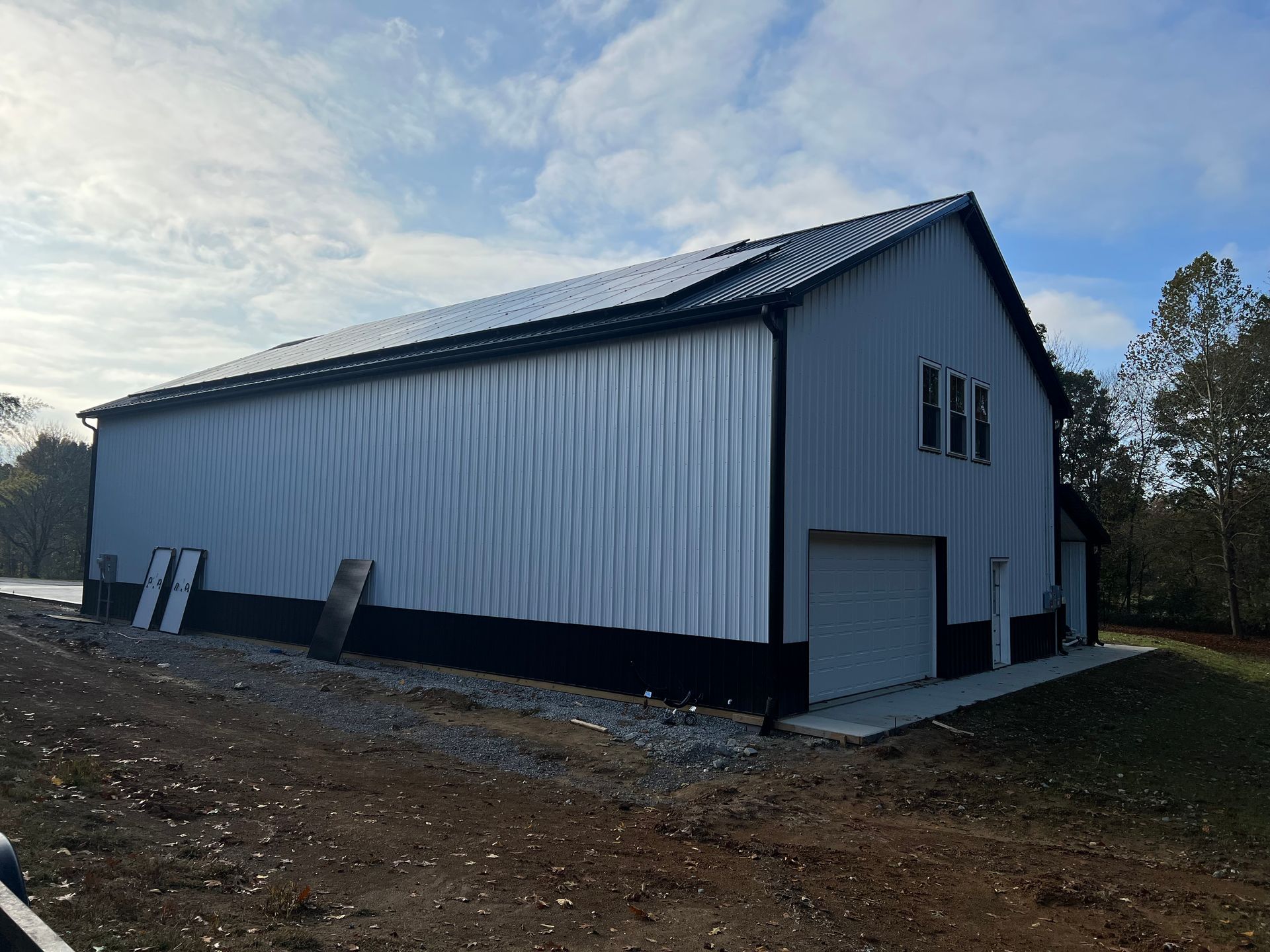A large, light gray metal barn with a black base, black trim, and a black roof with solar panels on a cloudy day. A concrete pad is in front of the garage door.