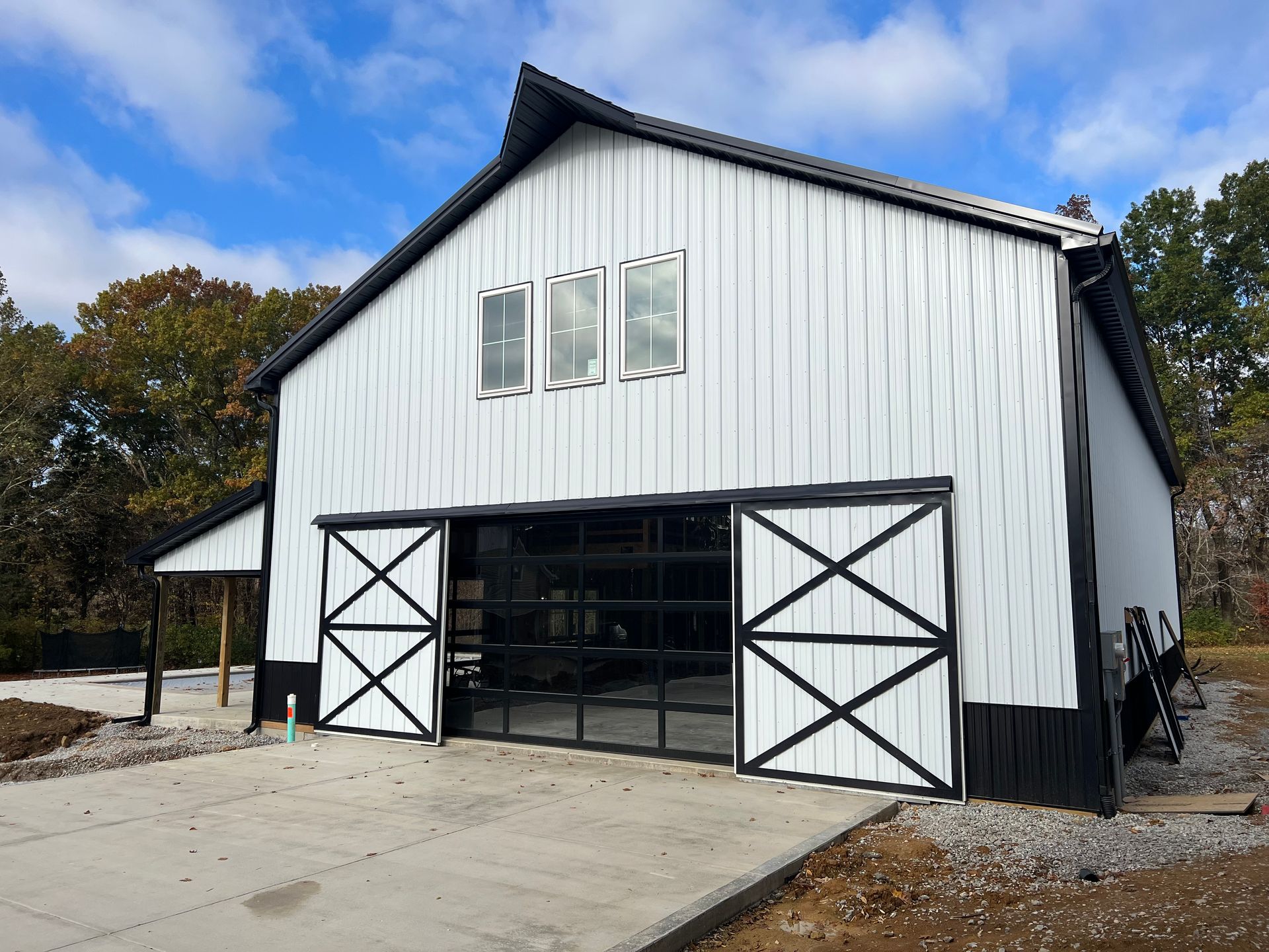 A modern barn with white siding, black trim, and a glass garage door stands against a cloudy blue sky. A concrete pad is in front.