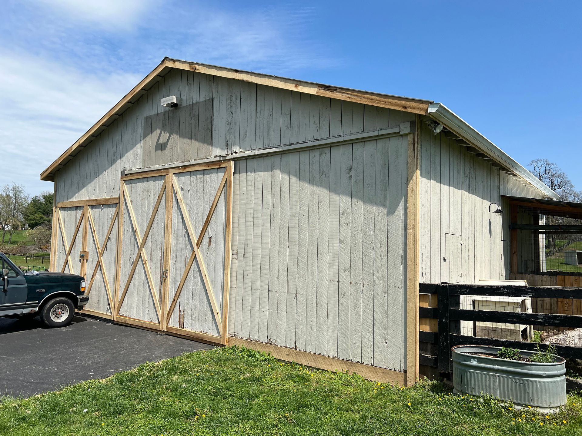 Weathered barn with large sliding doors; light gray siding, wooden trim, blue sky, and green grass.