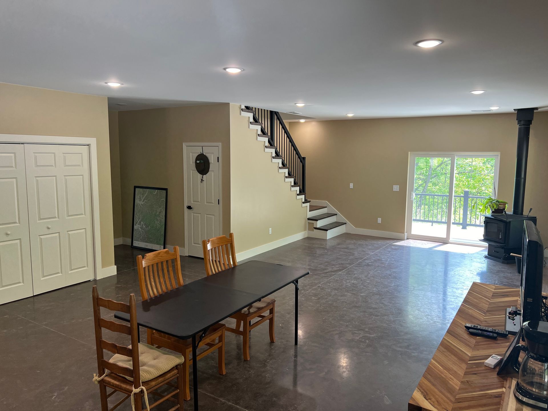 Spacious living area with stairs, a dark table and chairs, a wood stove, and a doorway to a balcony. Beige walls, gray flooring.