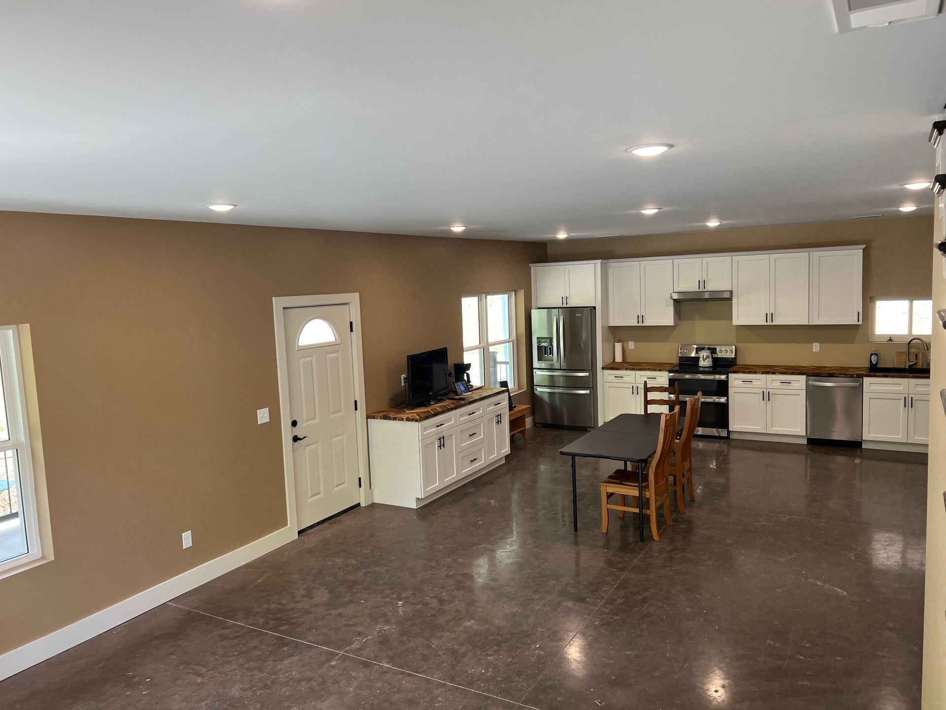 Open-plan kitchen with white cabinets, stainless steel appliances, and a long dark table, featuring brown walls and a concrete floor.