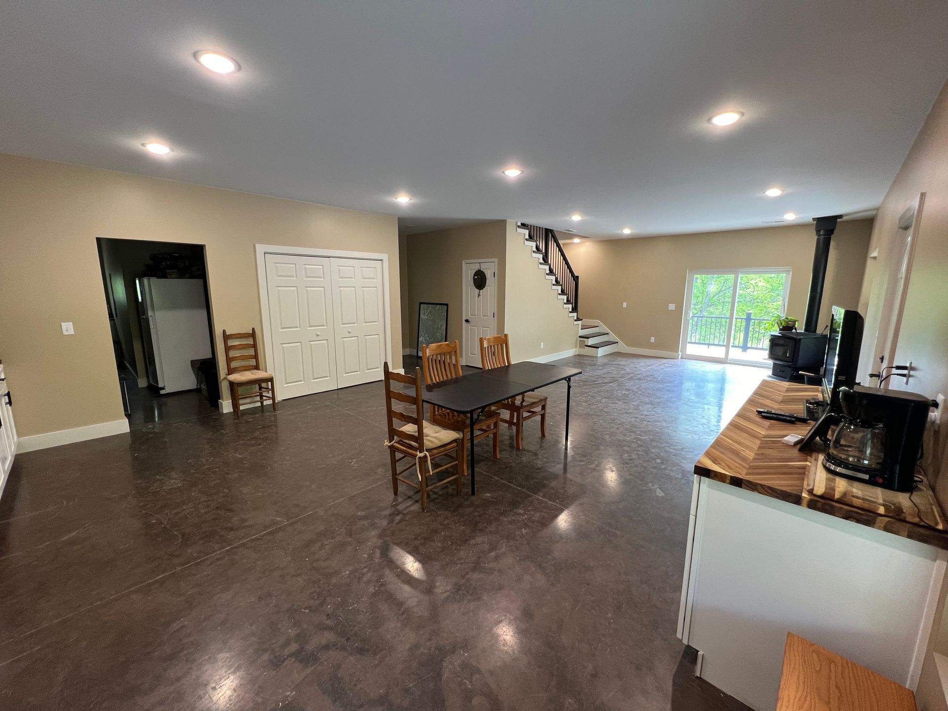 Spacious, open-plan interior with dark floors, tan walls, and recessed lighting. Includes a dining table, kitchen counter, and staircase.
