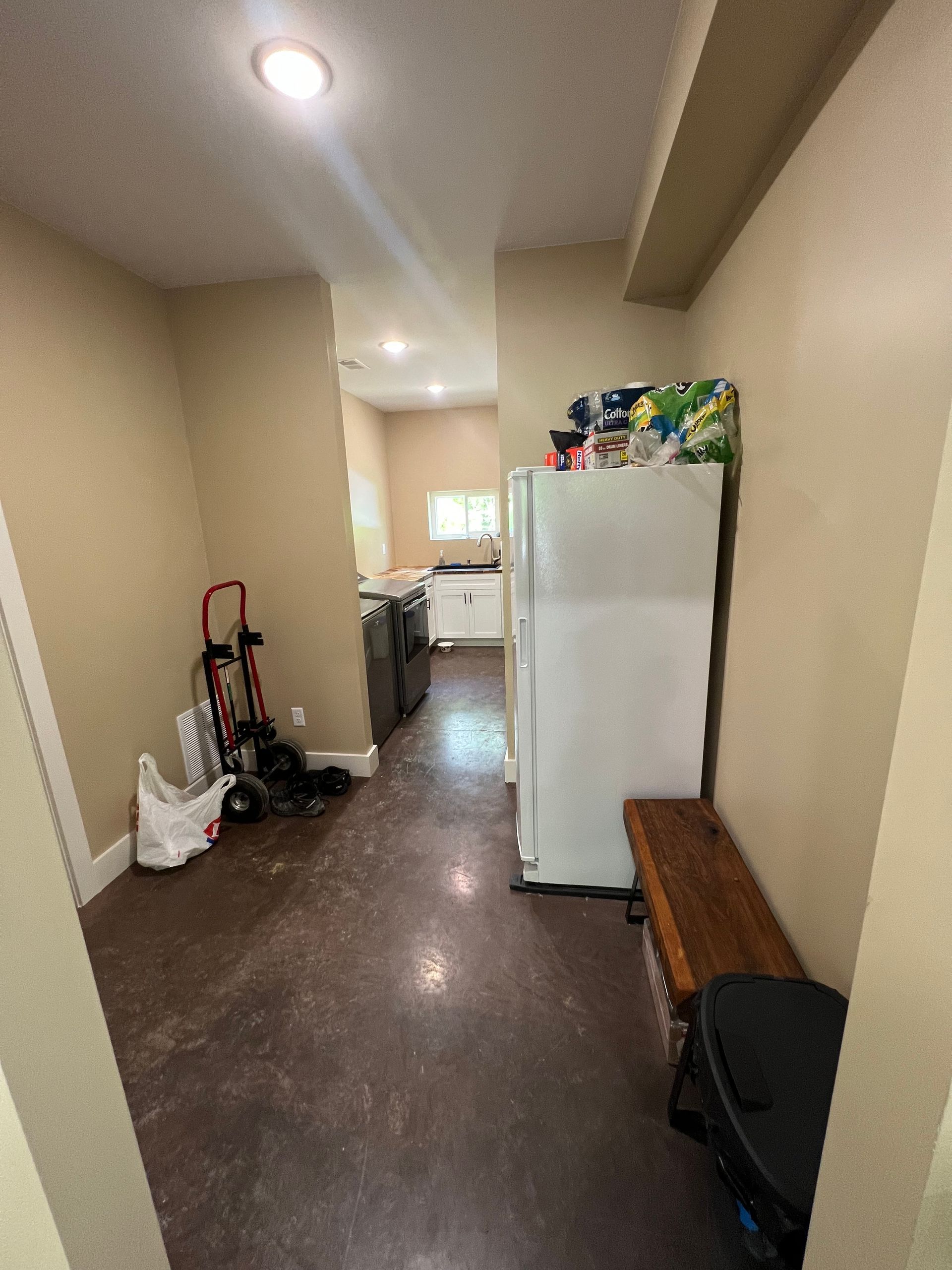Narrow hallway with a refrigerator, bench, and kitchen in the distance. The floor is dark, and walls are beige.
