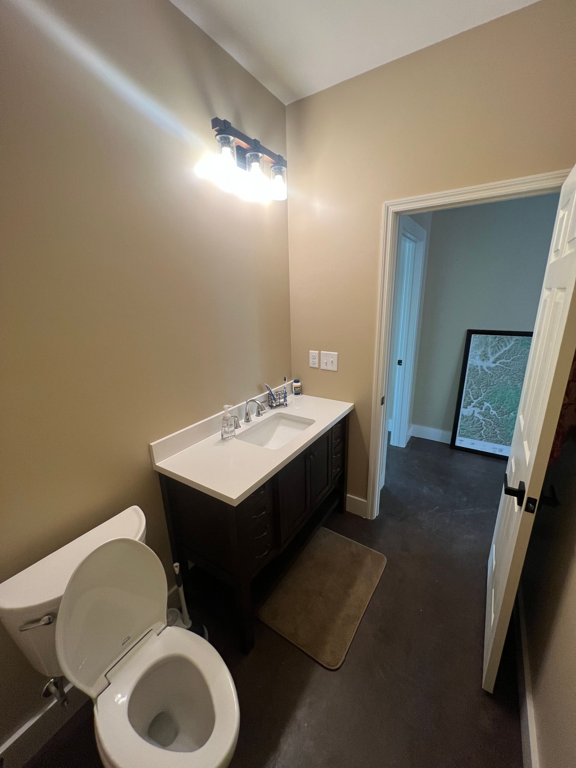 A small bathroom with a toilet, vanity, and doorway. The walls are tan, and the floor is dark.