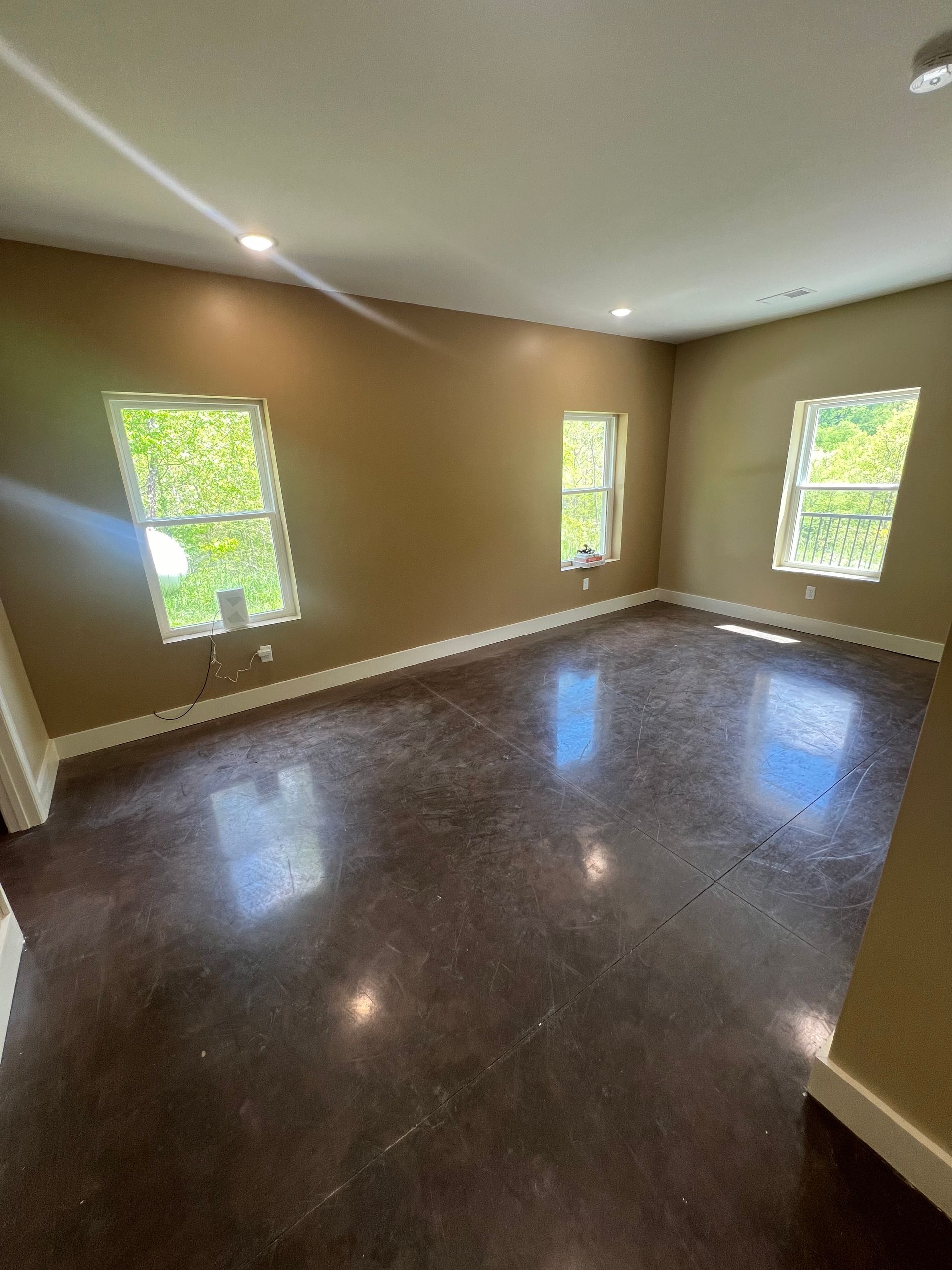 Empty room with brown polished concrete floor, three windows, and tan walls. White trim surrounds the windows and baseboards.