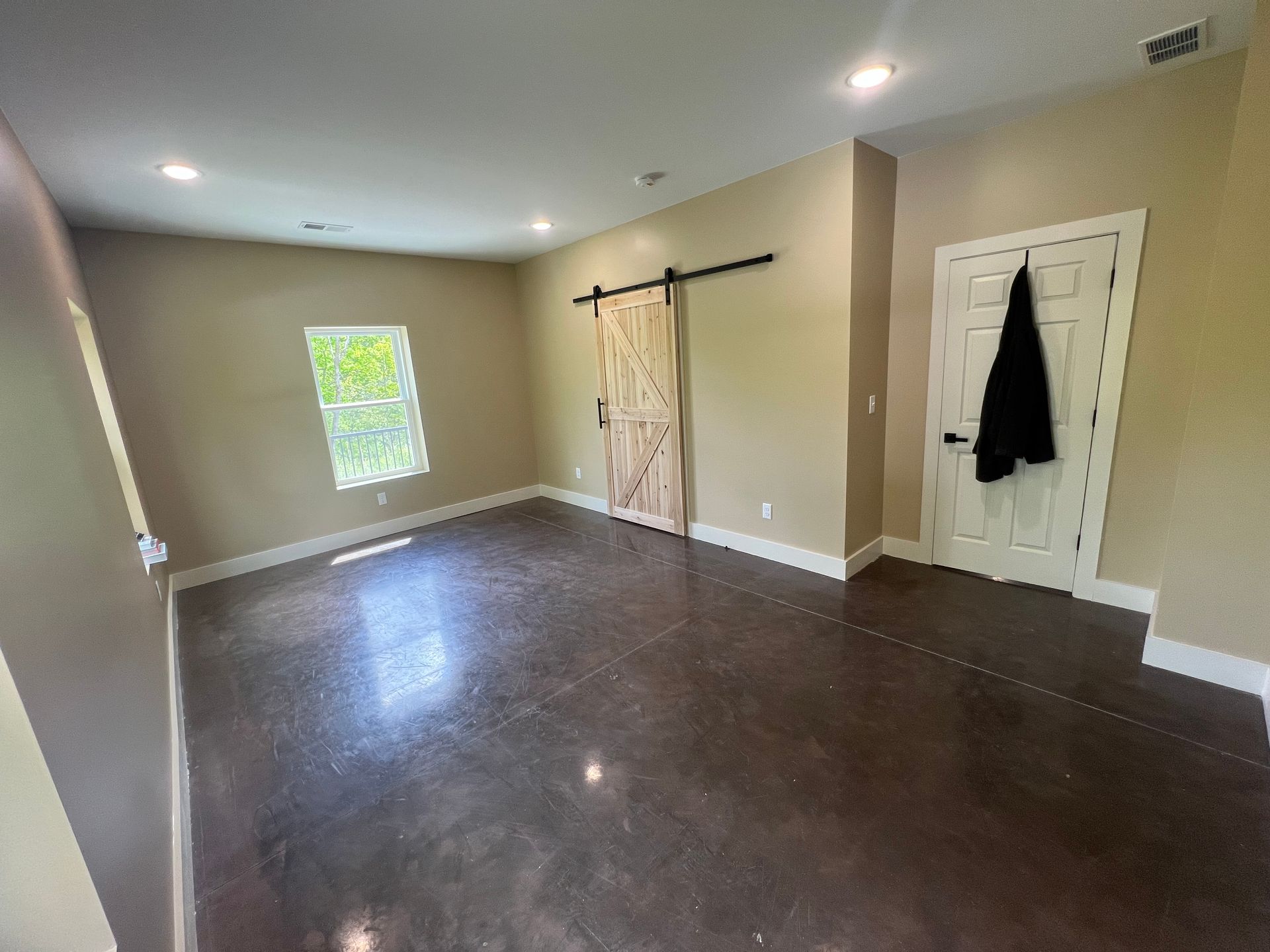 Empty room with dark gray flooring, light tan walls, and a wooden sliding door. A window and white door are visible.