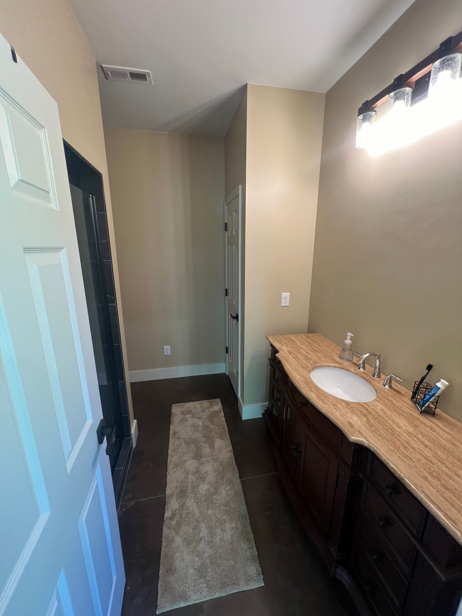 A narrow bathroom hallway with a dark wood vanity, tan countertop, and a rug on a dark tile floor.