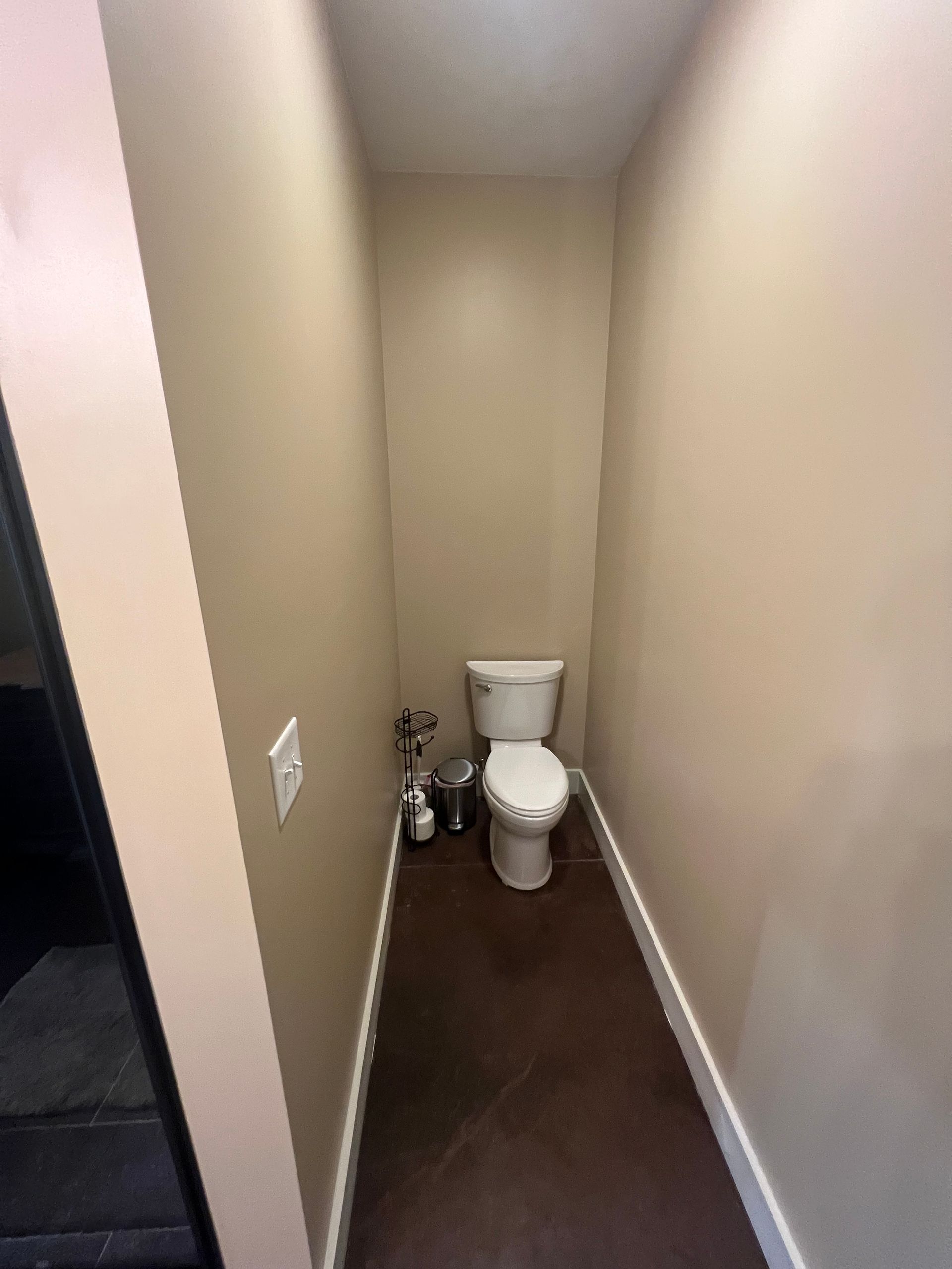 Small, narrow bathroom with a toilet, toilet paper holder, and trash can. Beige walls, dark brown floor, and white trim.