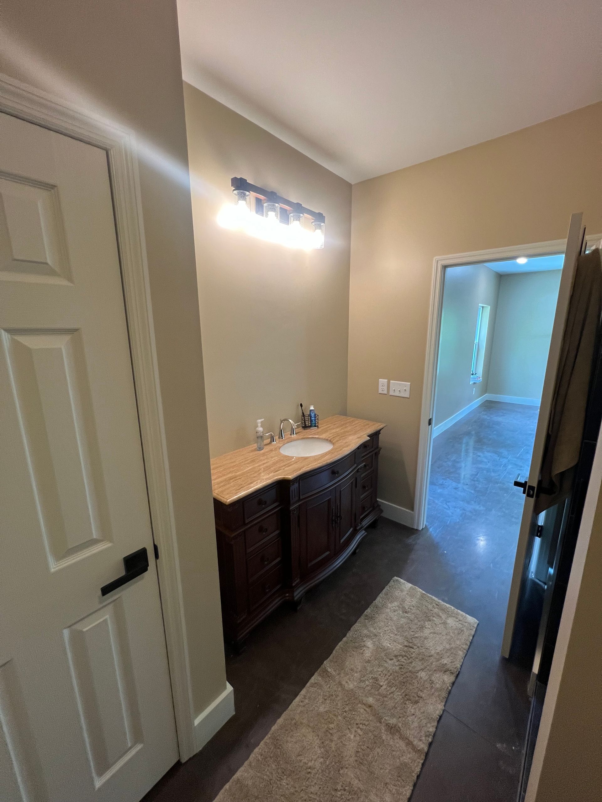 A small hallway with a vanity, rug, and open door. Dark wood vanity with a marble top, neutral walls, and dark flooring.