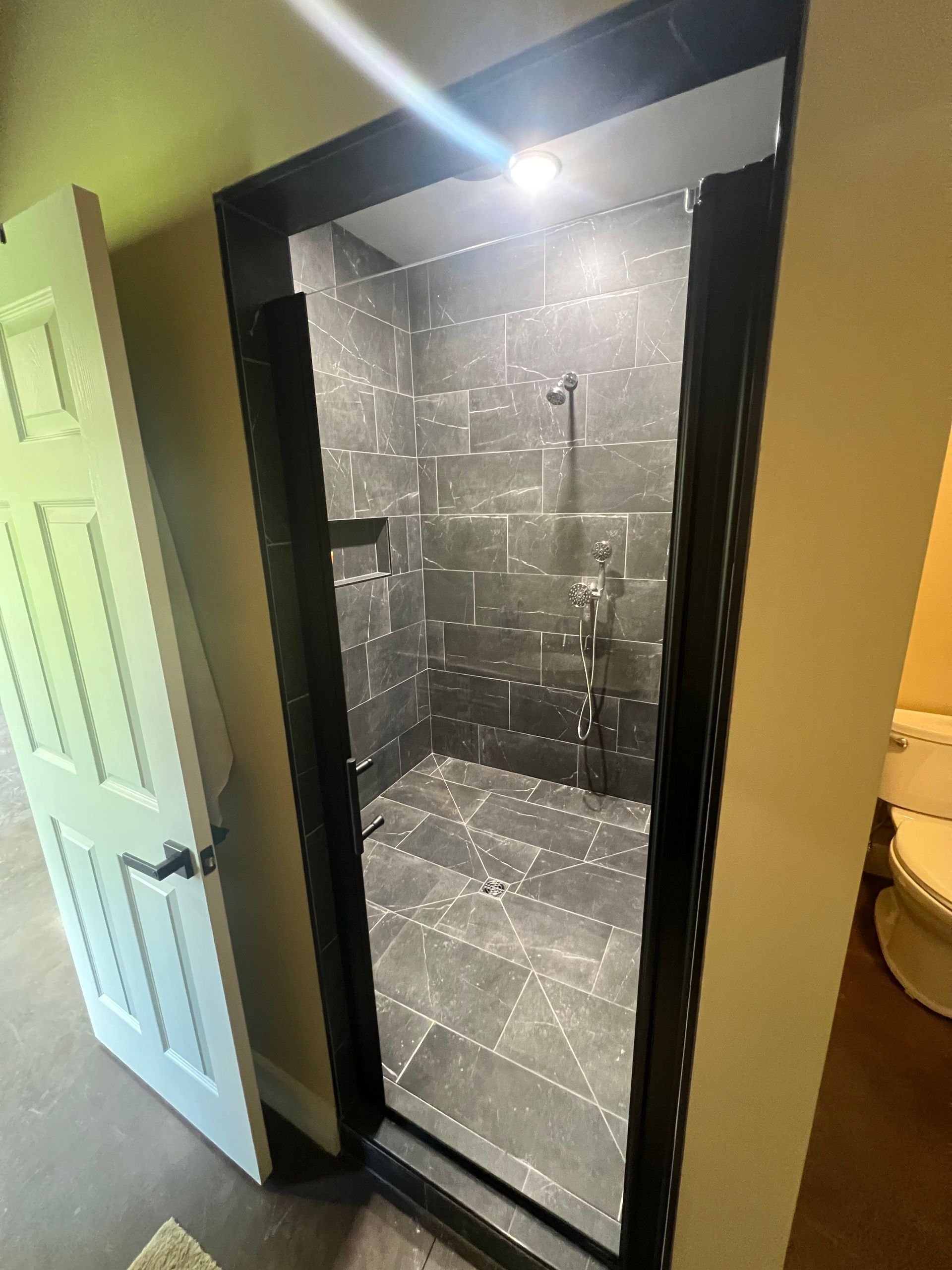 A dark-tiled shower with a glass door frame.  The shower is open, with a handheld showerhead and a small alcove.