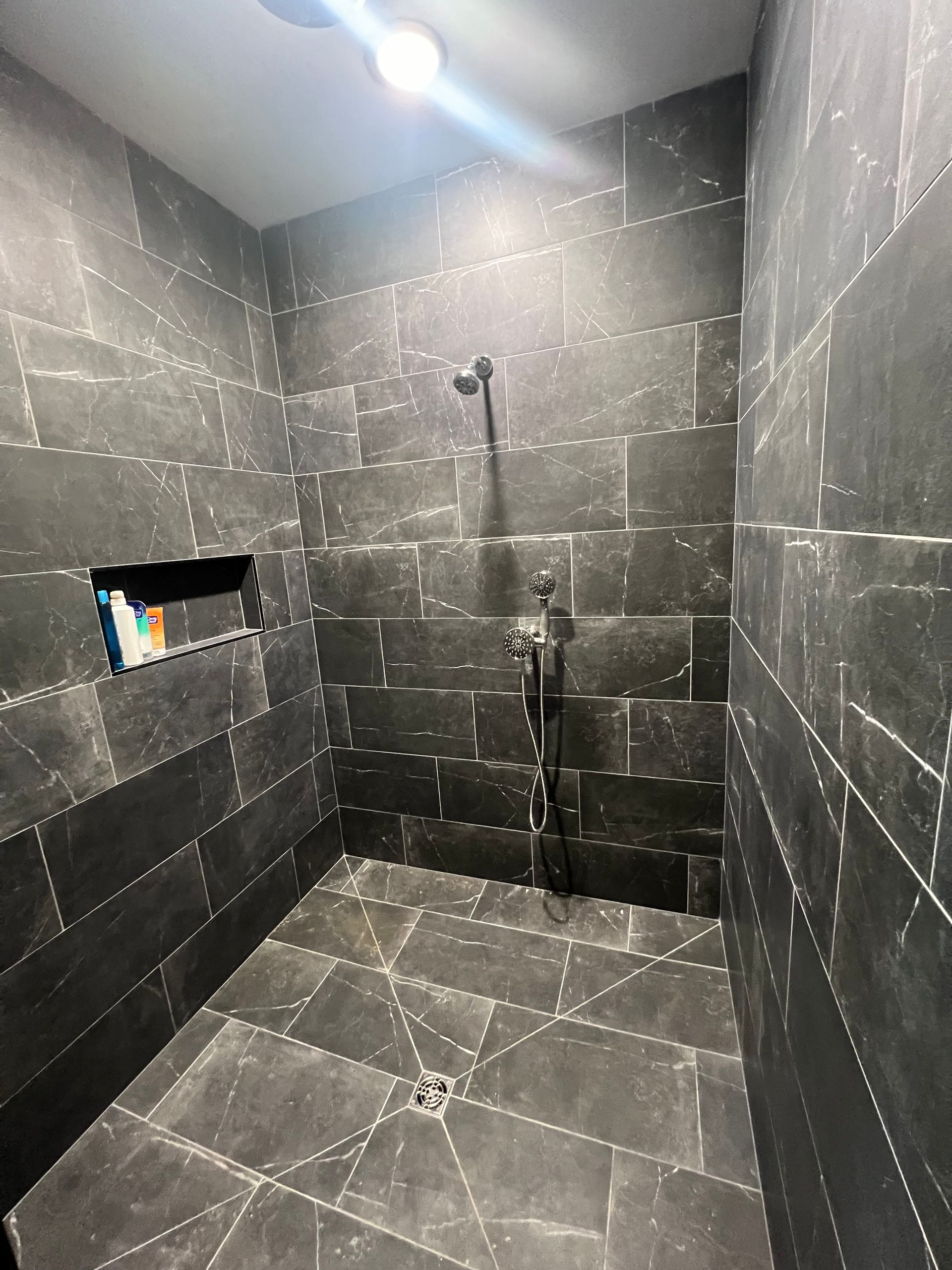 Dark gray tiled shower stall with a black shower head and a recessed shelf with product bottles.