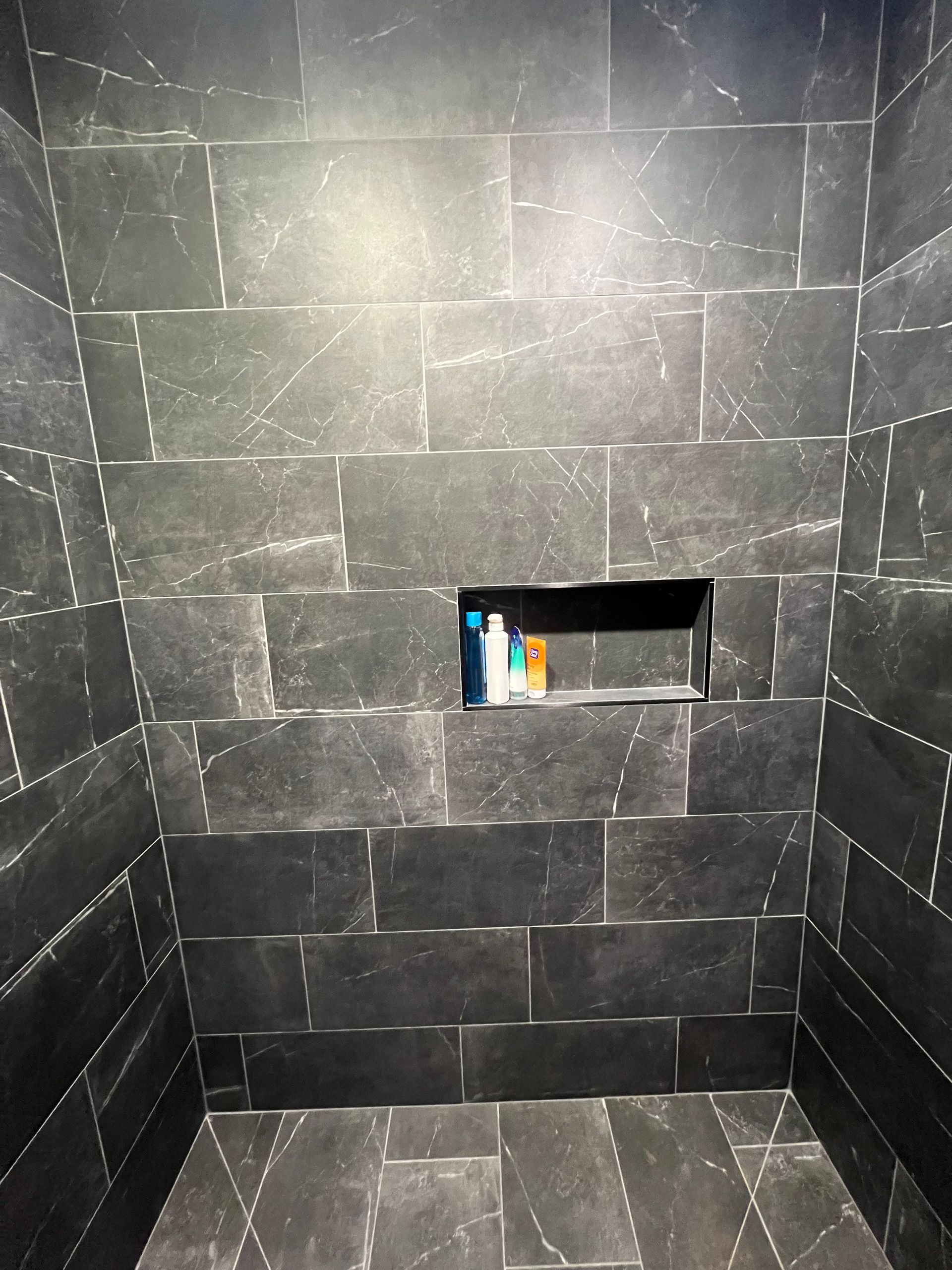 Dark gray tiled shower with a built-in shelf holding toiletries. Silver grout lines.