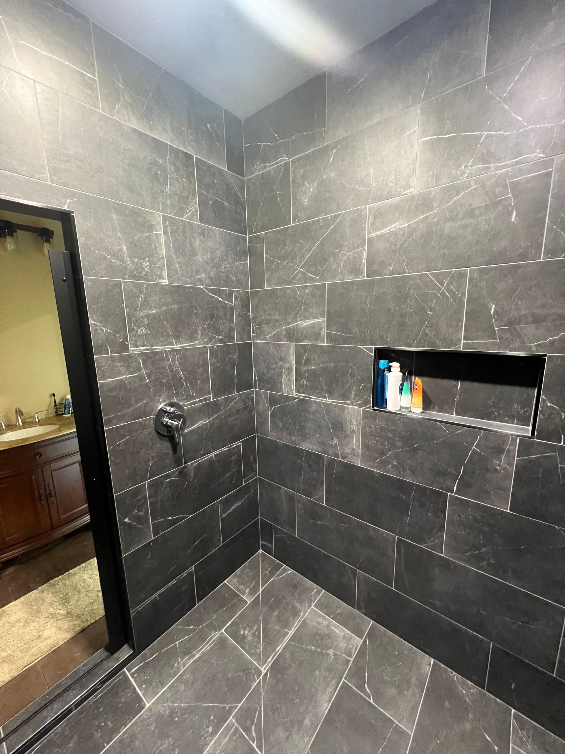 Dark tiled shower with a recessed shelf holding toiletries. The shower is open to a bathroom with a dark-framed door.