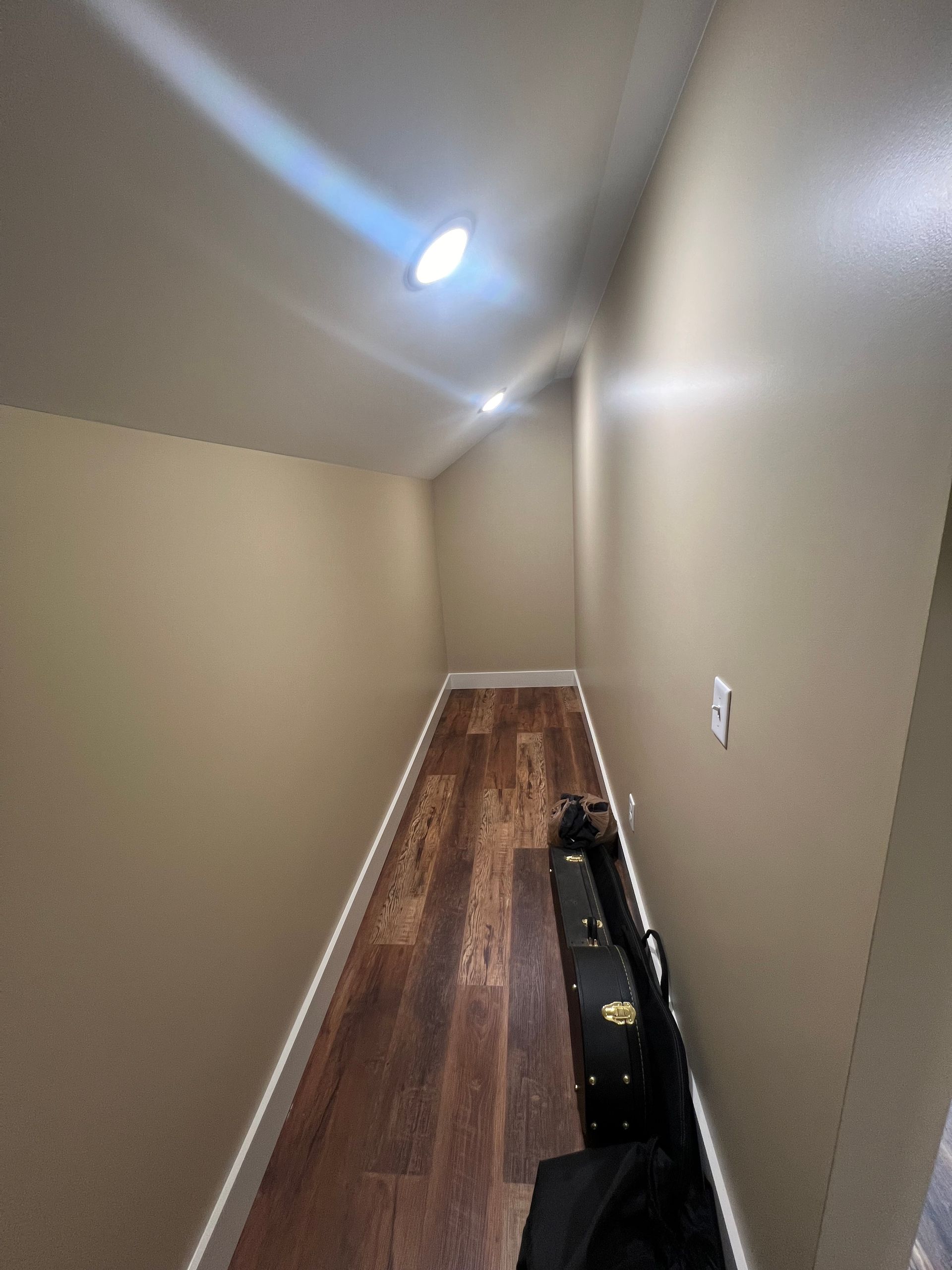 Narrow hallway with wood flooring, tan walls, and recessed lighting. A black case partially blocks the view.