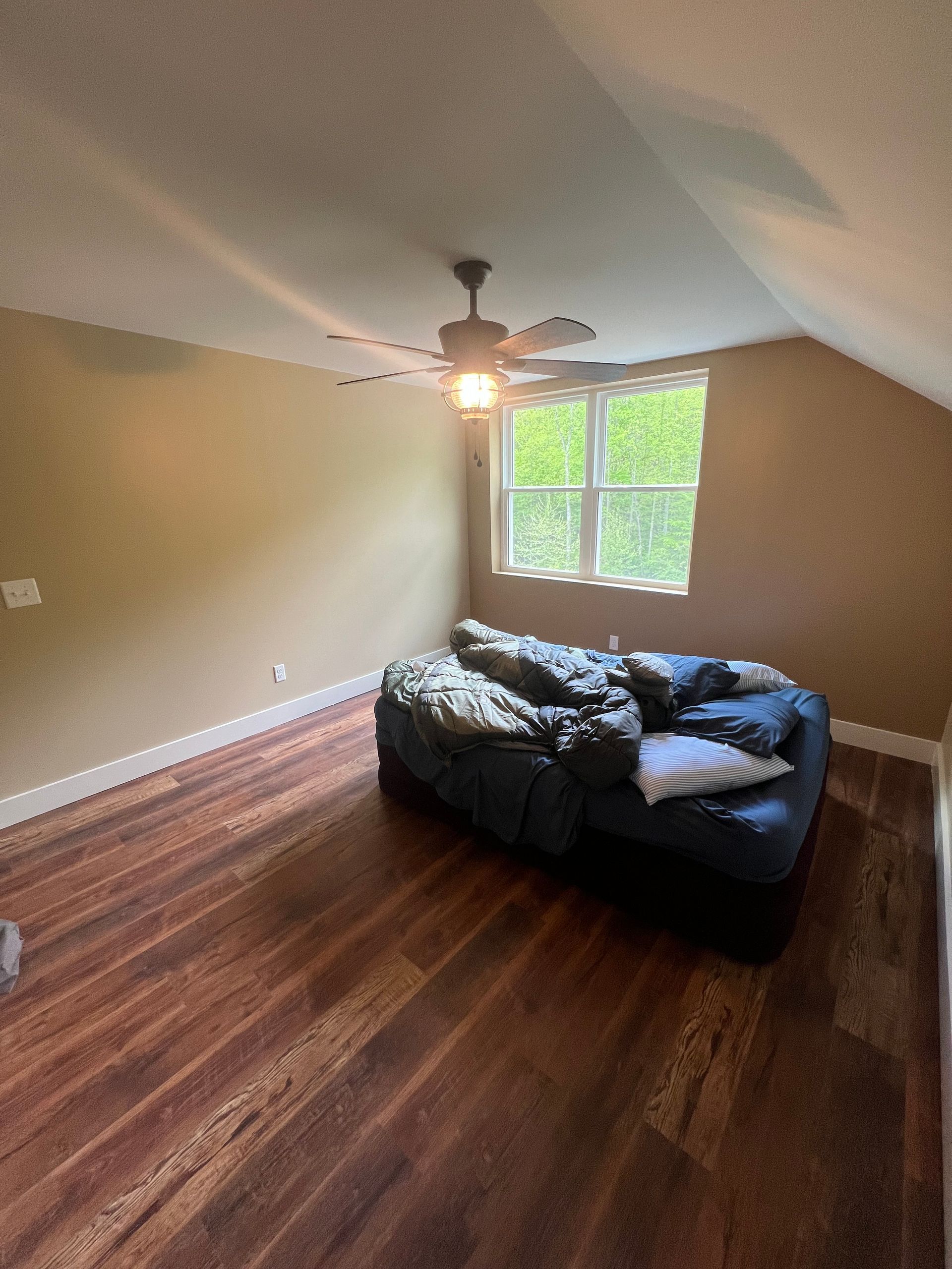 An attic bedroom with hardwood floors, a dark couch, and a window. The walls are tan. A ceiling fan is visible.