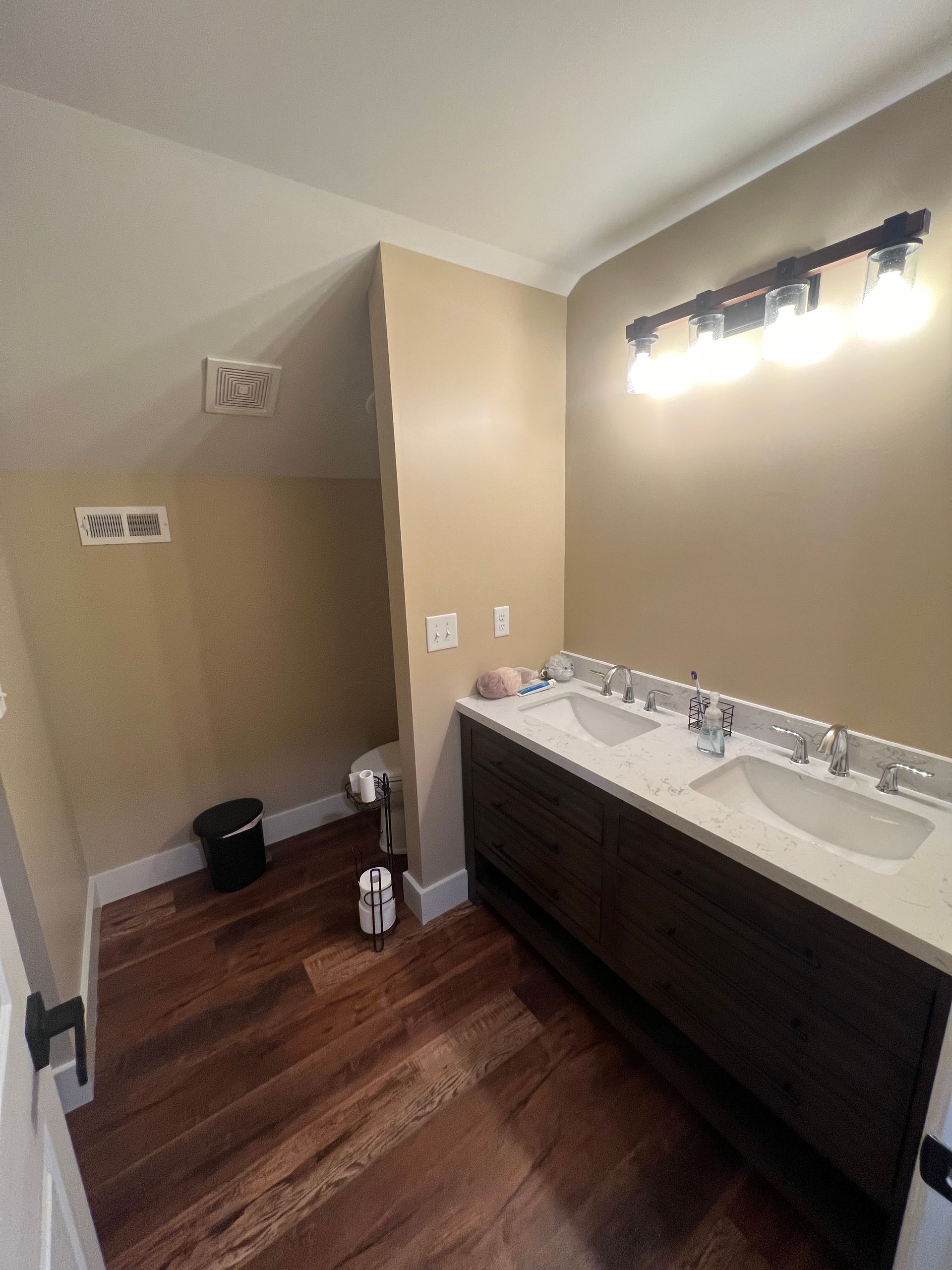 Bathroom with a double sink vanity, hardwood floors, and a light fixture. Dark wood vanity, white countertops, beige walls.